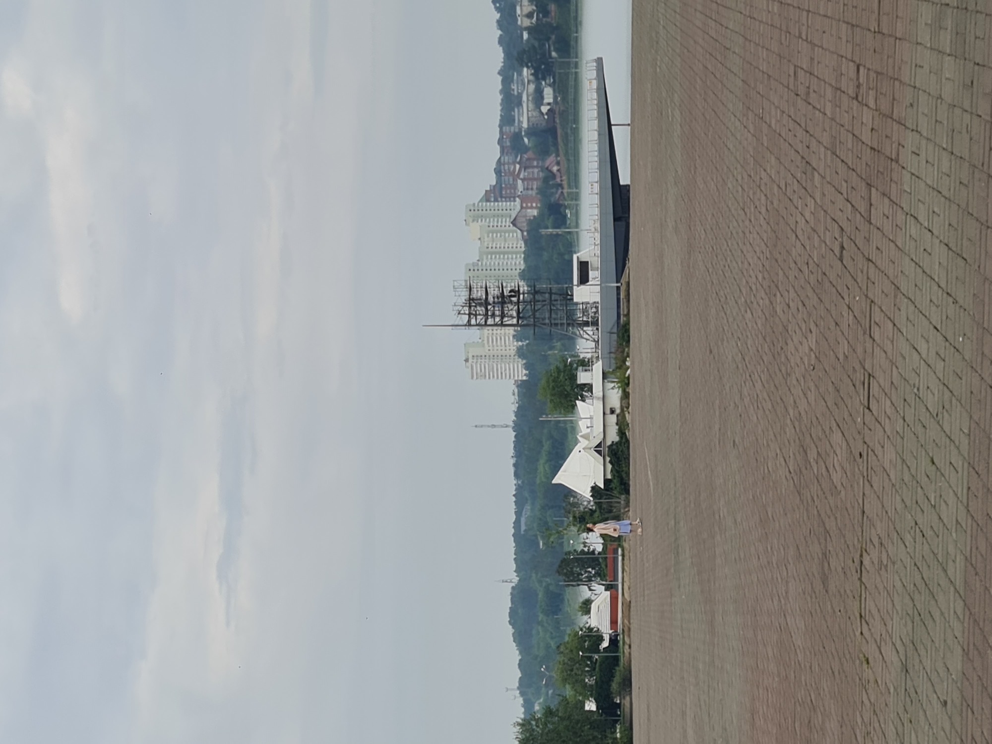 The image depicts a wide, paved area in front of a body of water, likely a river or lake. In the foreground, the ground is covered with reddish-brown brick pavers. A single woman, wearing a light-colored dress or skirt and a light brown jacket, is walking across the paved area from left to right.
In the mid-ground, there are several buildings. To the left, there are smaller, white buildings with red roofs, and some trees. In the center, a distinctive modern white building with a triangular roof structure is visible. Adjacent to this, and extending towards the water, is a grey, platform-like structure that appears to be under construction or renovation, with scaffolding and metal frameworks erected on top of it.
Across the water, on the opposite bank, a cityscape is visible with numerous multi-story apartment buildings, some light-colored and modern, others with red brick. Green trees and hills form the backdrop to the urban landscape. The sky is overcast with grey clouds.
This scene is located at the Angara Embankment (Naberezhnaya Angary) in Irkutsk, Russia. The structure under construction with scaffolding is likely part of the reconstruction or development of the embankment area, potentially related to the "Strelka" (Arrow) development which is a prominent landmark in Irkutsk at the confluence of the Angara River and the Irkut River. The large paved area is a public space for recreation and events.