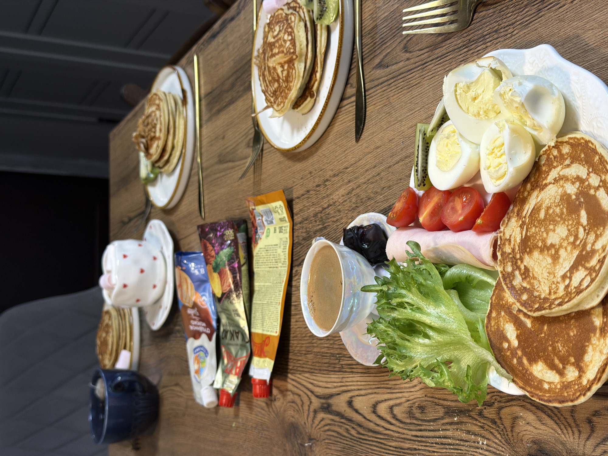 The image shows a wooden table laden with breakfast items, possibly in a home or apartment setting. There are three plates of pancakes with kiwi slices, each with a knife and fork placed nearby. A fourth plate features pancakes, boiled eggs, tomato slices, ham slices, lettuce, and black plums. A cup of coffee rests on a saucer near the edge of the table. To the left of the coffee are tubes of what appear to be condensed milk with cocoa flavorings. Two additional cups sit stacked beneath a pancake plate, near the table's edge. While the specific location in Perm, Russia cannot be determined from the image alone, the presence of food items and common household objects indicates an interior setting.