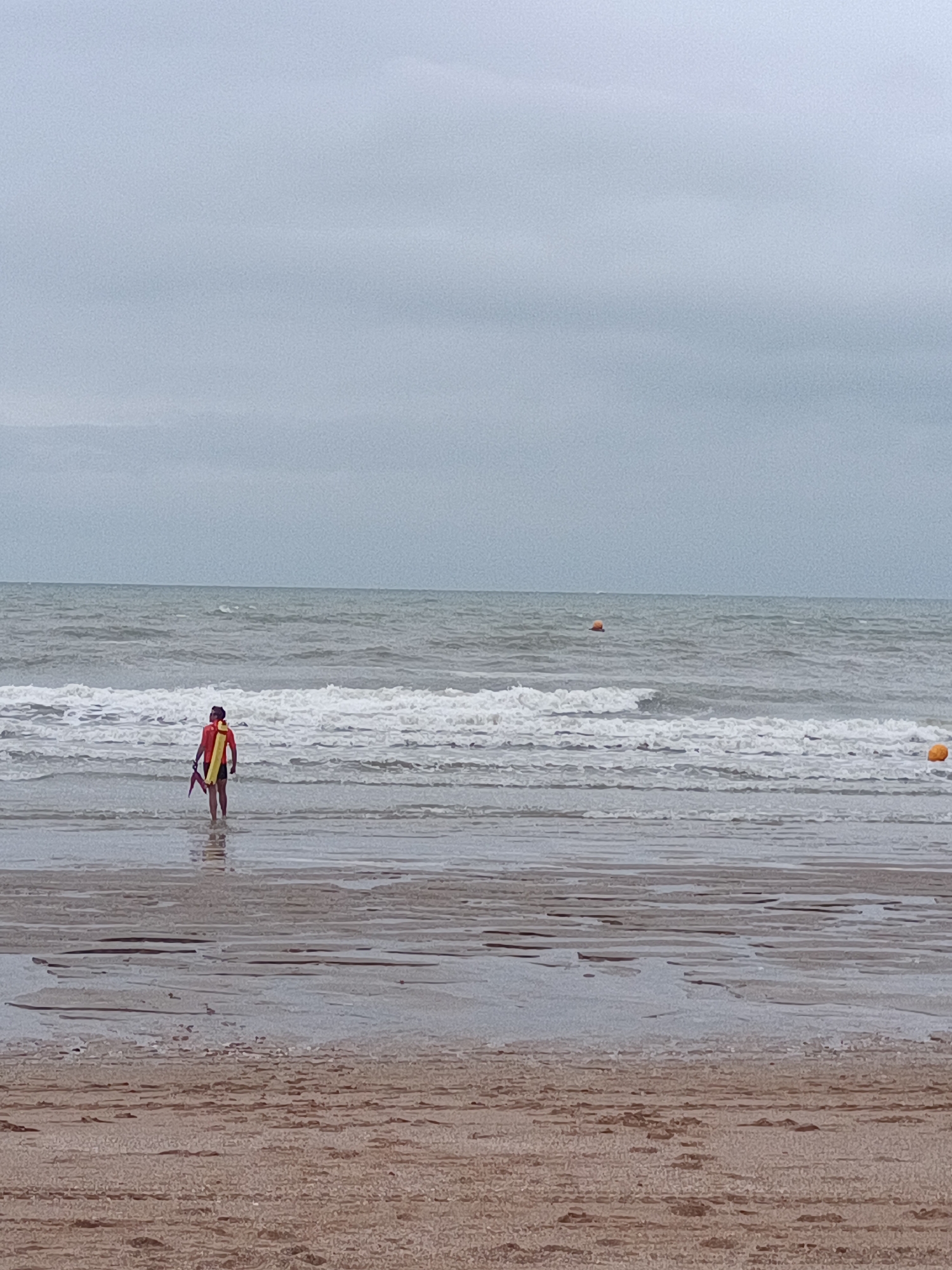 A solitary figure, possibly a lifeguard, stands in the shallow water at the edge of the sea. They are wearing a bright orange vest and are holding a red and yellow object, which could be a rescue buoy or a signaling device. The person is facing away from the camera, towards the ocean, which is characterized by choppy waves under an overcast sky. Several orange buoys are visible in the water, indicating a designated swimming or boating area. The beach itself is sandy with visible patterns left by receding water, suggesting a tidal shore. The overall scene depicts a calm but potentially challenging coastal environment. This appears to be a public beach in Houlgate, France, given the typical appearance of French coastal towns.