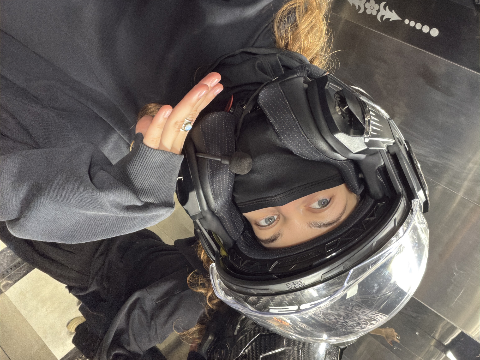 A high-angle portrait shot shows a person looking up at the camera, wearing a dark motorcycle helmet and a black balaclava. The person's face is mostly covered by the balaclava, but their light-colored eyes and eyebrows are clearly visible, framed by the helmet visor. Long, curly brown hair spills out from under the helmet.
The person is wearing a dark grey or black hooded sweatshirt. Their left hand is raised slightly, revealing light pink nail polish and a silver ring with a light blue, possibly opal-like, stone on their ring finger. A small black microphone from an integrated communication system is positioned near their mouth, attached to the helmet's interior padding.
The helmet itself is dark, with reflective silver-colored accents on the sides. The clear visor shows some reflections, including possibly the person's other hand or part of a vehicle. The brand "LS2" is faintly visible on the visor.
The background consists of a dark, reflective surface, possibly a polished floor or wall, with a decorative white floral/geometric pattern on it. Below the person's mid-section, a tiled floor with light and dark squares is visible, suggesting an indoor setting like a showroom or a garage. The person appears to be seated or leaning.