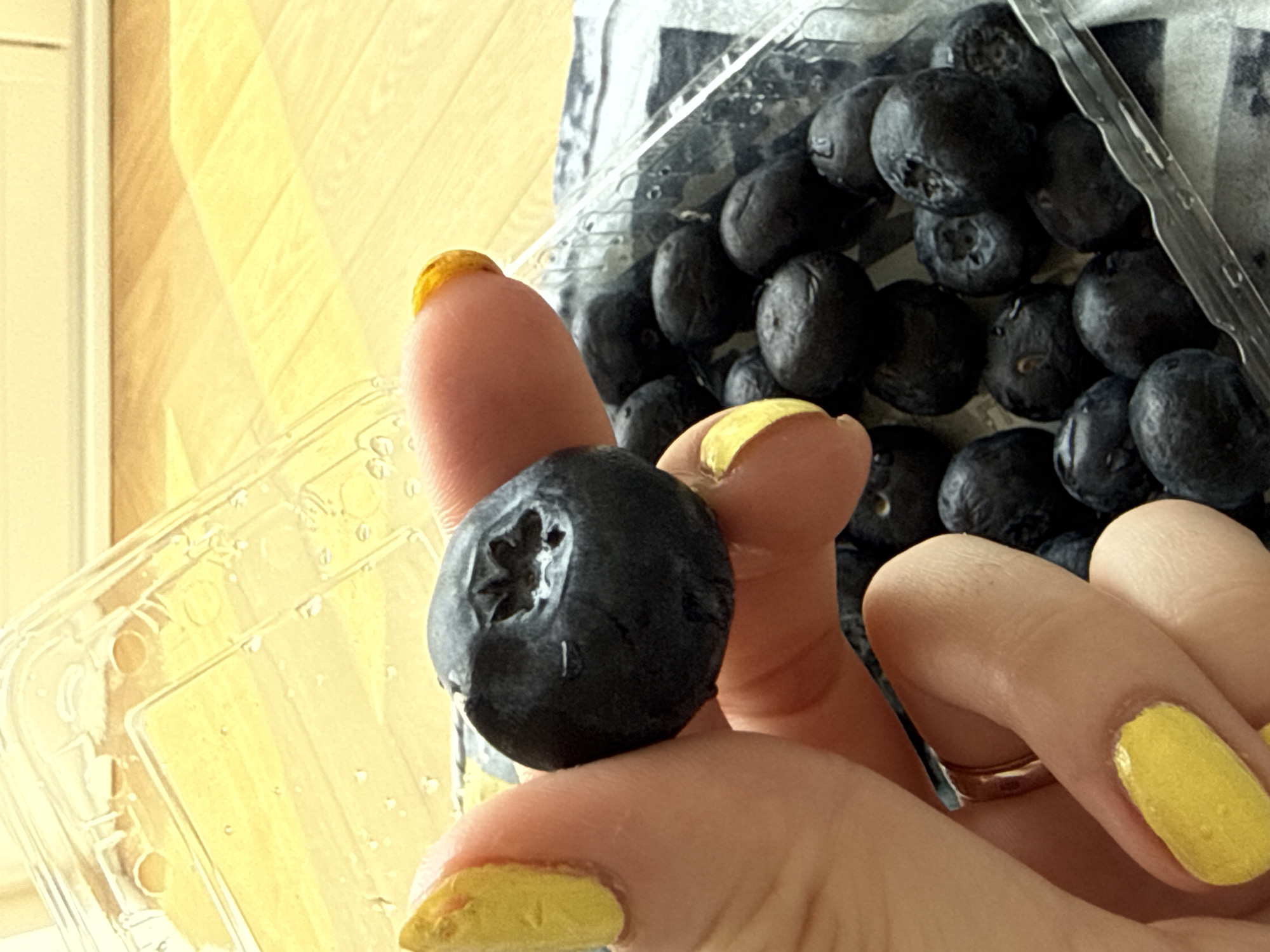 A close-up shot depicts a hand holding a single blueberry, with a container of more blueberries visible in the background. The fingernails are painted yellow. The image appears to be taken indoors. The location is not discernible from the image.
