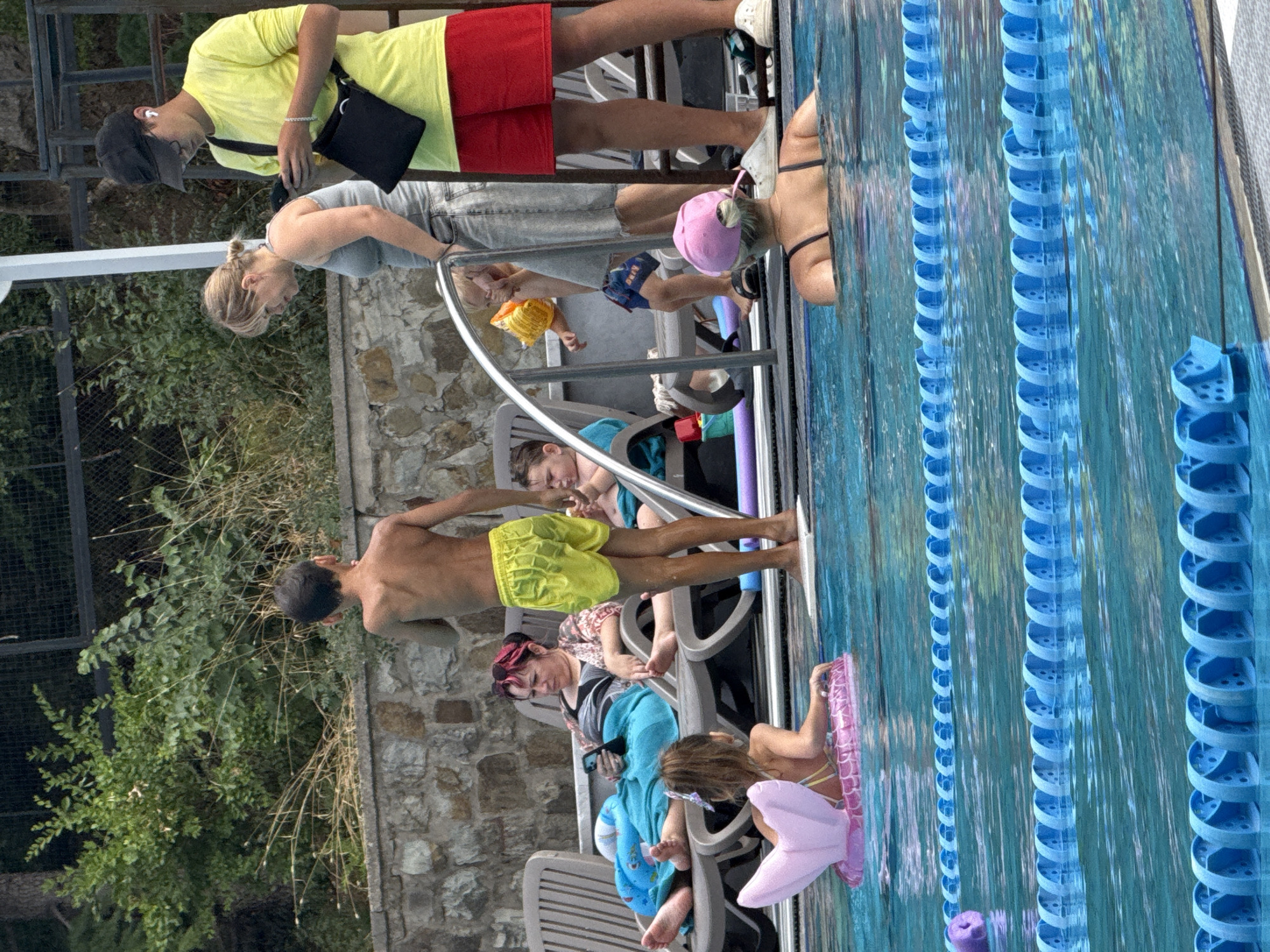 In the city of Yalta, Ukraine, a group of people are gathered around a swimming pool on a sunny day. In the foreground, a young girl with dark hair, wearing a pink mermaid-themed inflatable ring, floats in the pool. Behind her, a woman with blonde hair in a bun, wearing a grey tank top and denim shorts, is partially submerged in the water.
On the poolside, several people are relaxing on lounge chairs. A boy with dark hair, wearing yellow swim trunks, stands near the pool edge, facing away from the camera. To his left, a woman with dark hair and pink highlights is seated on a lounge chair, looking at her phone. Beside her, two children are visible. One, a boy, is wearing a yellow flotation device. Another child, wearing a blue towel, is also seated.
Further back, a young man wearing a yellow t-shirt, red shorts, a black baseball cap, and a black bag over his shoulder is looking at his phone. The environment includes a stone wall, lush green bushes, and a fence in the background. The pool has blue lane dividers.