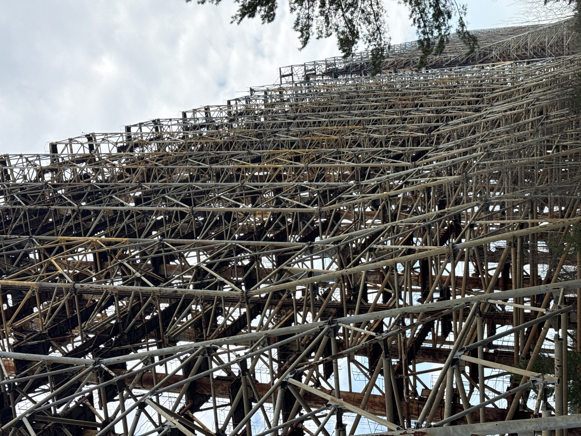The image shows the Duga radar system, located in Chornobyl, Ukraine. The structure is a massive, steel lattice framework that rises steeply toward the sky. The latticework is complex and repetitive, with a combination of horizontal, vertical, and diagonal supports. The steel appears weathered and rusty in sections, and some parts are blackened. The sky is overcast, with cloudy conditions. Tree branches are visible in the top corners.