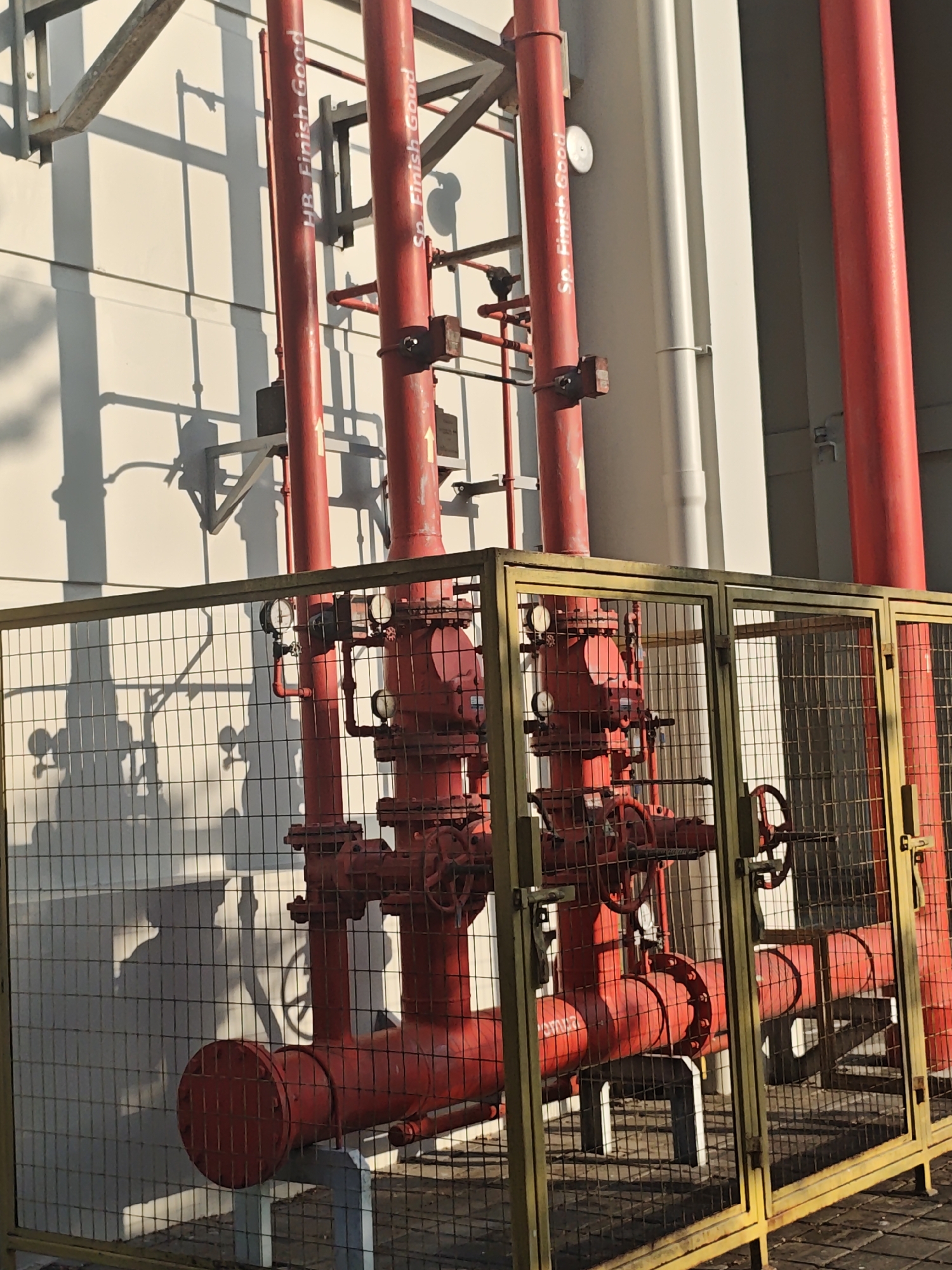 The image depicts a section of an industrial facility, specifically focusing on a network of bright red pipes, likely part of a fire suppression system. These pipes are enclosed within a yellow-painted metal cage with wire mesh fencing. Several pressure gauges are visible on the pipes, indicating operational status. The pipes are mounted on concrete supports. In the background, a light-colored wall is illuminated by sunlight, casting prominent shadows of the pipes and associated structures onto the wall. The overall scene suggests an outdoor industrial setting, potentially within Bae, Indonesia, given the context. There are no people visible in the image.
