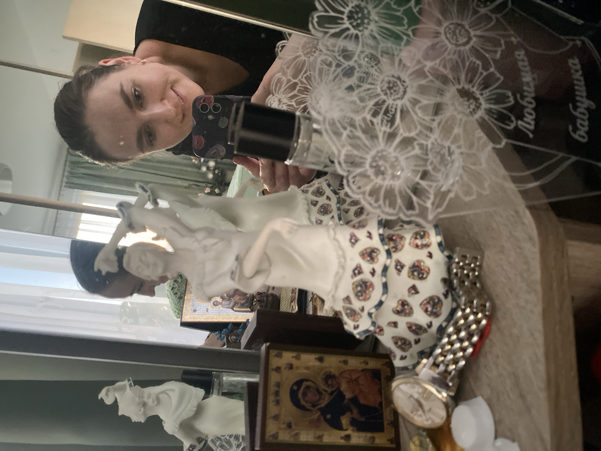A woman is taking a selfie in a mirror, reflecting a shelf of various items. The shelf displays religious icons, decorative figurines, and a watch. In the mirror's reflection, a woman is seen holding a smartphone. Etched on the glass near the upper right corner are words that translate to "beloved grandmother."
The exact location within the city of Yekaterinburg, Russia cannot be conclusively determined from the given image.