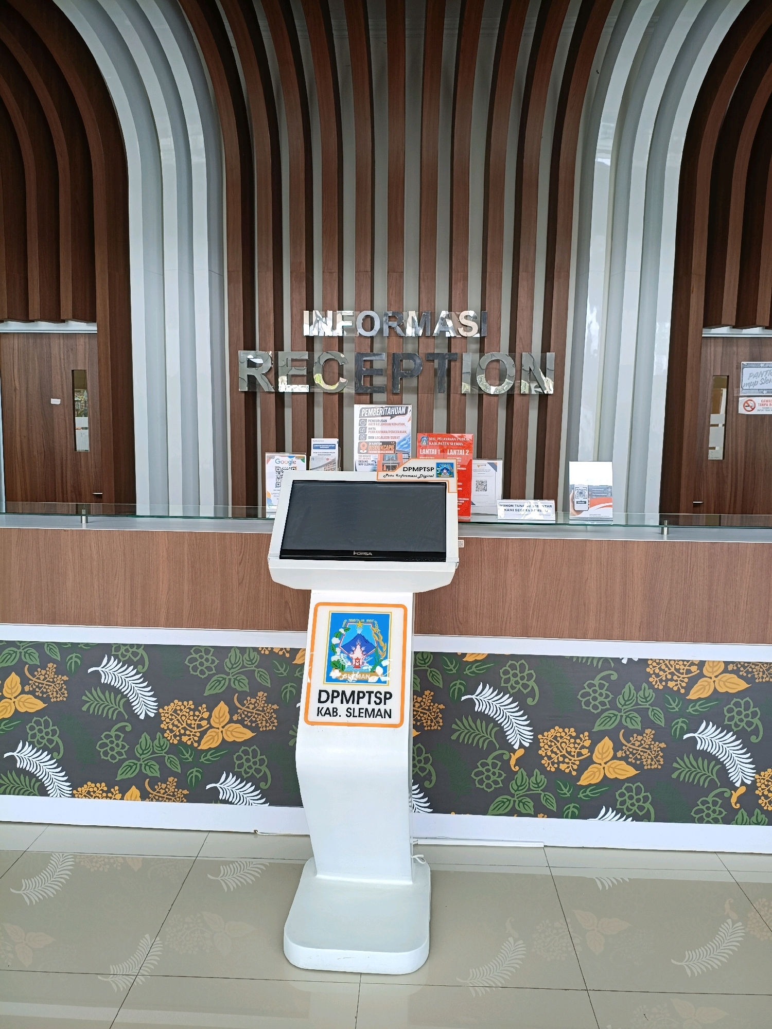 A white kiosk with a touch screen display is positioned in front of a reception desk. The kiosk displays the text "DPMPTSP KAB. SLEMAN" and a logo. Behind the reception desk, metallic letters spell out "INFORMASI RECEPTION". The reception desk itself is made of wood with a glass counter. The background features a wall with curved wooden and white vertical panels. On the reception counter, several informational pamphlets and signs are visible. The floor is tiled with a light beige color. The overall environment appears to be an indoor public space, likely a government office or information center within Sleman, Indonesia.