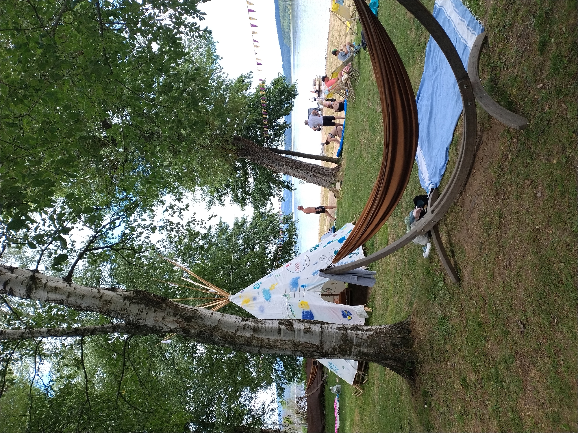 The image depicts a lakeside scene in Radslav, Czechia, on a grassy area adjacent to a sandy beach. In the foreground, a brown hammock is suspended from a wooden stand, draped over a blue and white striped towel. Behind the hammock, a white teepee decorated with colorful drawings stands tall. Several trees with green foliage are scattered throughout the scene, providing shade. In the background, people are visible near the water, some sitting on lounge chairs and others walking along the shore. Boats are present in the lake, and a string of purple and yellow flags is strung between trees along the beach. The overall impression is one of a relaxed outdoor gathering or camping event by the water.