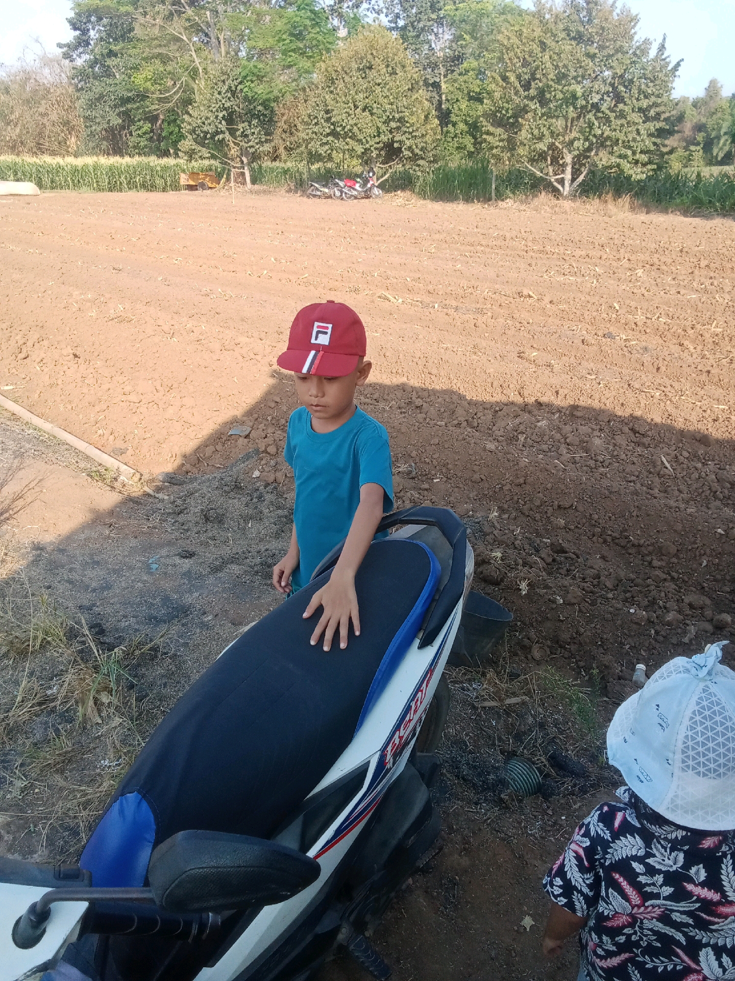 In an outdoor scene in the city of Unknown, Indonesia, a young boy wearing a red baseball cap with a white "F" logo and a blue t-shirt stands next to a motorcycle. The motorcycle has a dark blue seat cover with a blue accent. In the background, a field of dry earth is visible, with trees and foliage beyond. A second child, wearing a white patterned hat and a dark floral shirt, is also present, facing away from the camera. The ground appears to be a mix of dirt and dry grass.