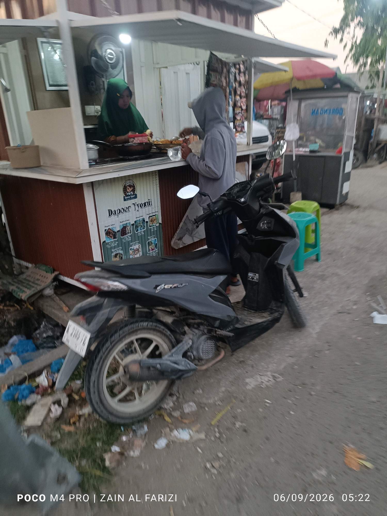 A blurred, full shot captures an outdoor food stall in Arosbaya, Indonesia, at dusk. A woman wearing a green hijab is preparing food behind the counter, illuminated by a bright light. A person in a grey hooded sweatshirt stands beside her, facing away from the camera. A parked black motorcycle is in the foreground, with its license plate partially visible. In the background, a colorful umbrella shades part of the stall, and a refrigerator unit with writing on it is visible. The ground is covered in dirt and debris, and several small green plastic stools are scattered around. The overall impression is that of a busy, informal market setting.