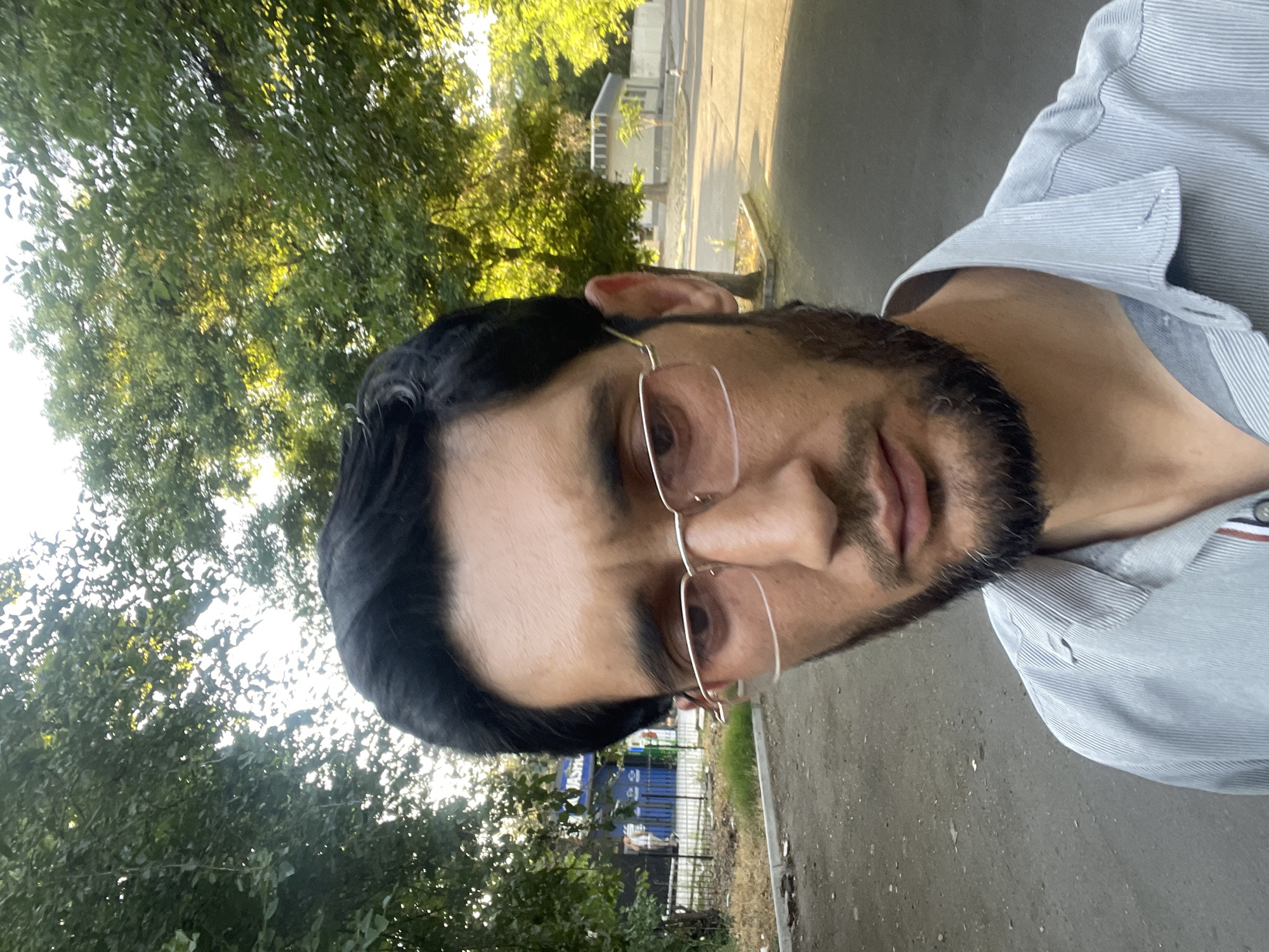 This is a selfie of a man wearing glasses and a light-colored striped shirt, standing outdoors in Tashkent, Uzbekistan. He has dark hair and a beard. Behind him, trees with green foliage are visible, and further in the background, there is a street, a sidewalk, a building with a blue facade, and some fencing. The lighting suggests it is daytime, possibly late afternoon, with sunlight filtering through the trees.