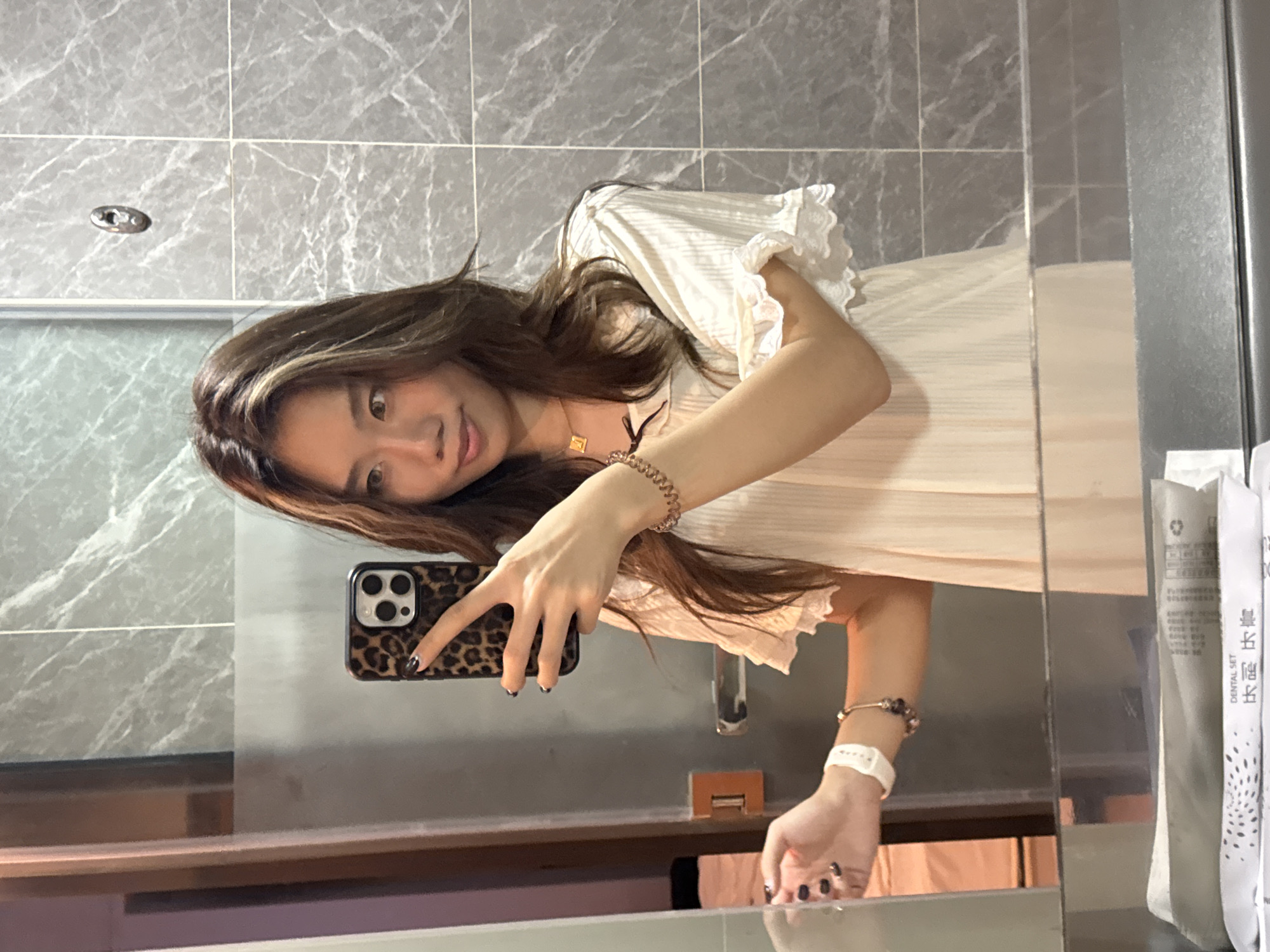 The image shows a woman taking a selfie in what appears to be a bathroom. She is holding a phone with a leopard print case in her right hand. She's wearing a light-colored dress with frills and bracelets on both wrists. The bathroom has gray marble tiles. Dental kits are visible near the edge of the mirror. There's no specific information in the image to confirm that the location is Chenghua District, China.
