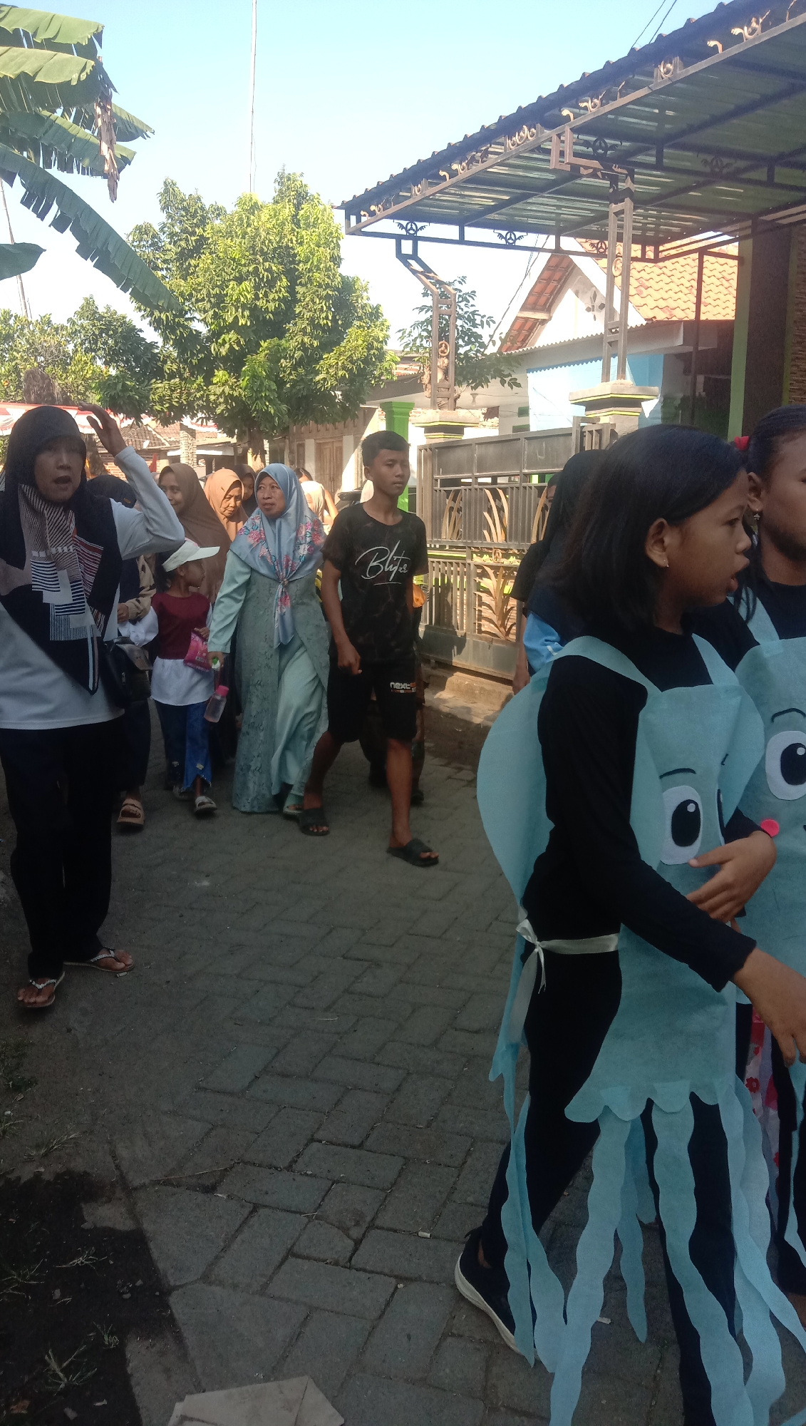 A group of people, including adults and children, are walking down a paved street under a clear sky, with trees and buildings visible in the background. The street is lined with patterned paving stones. In the foreground, two young girls are wearing teal costumes resembling jellyfish, with large cartoon eyes. They are walking towards the right of the frame. To their left, a teenage boy in a black t-shirt and shorts is walking. Further left, a woman in a light blue hijab and a long dress walks alongside a younger child in a red shirt and blue pants. Another woman in a black headscarf is on the far left, gesturing with her hand. Behind them, other individuals are walking, some partially visible. A covered awning with decorative metalwork extends over a walkway to the right of the street. The scene appears to be a daytime procession or parade on a street in Ngempit, Indonesia.