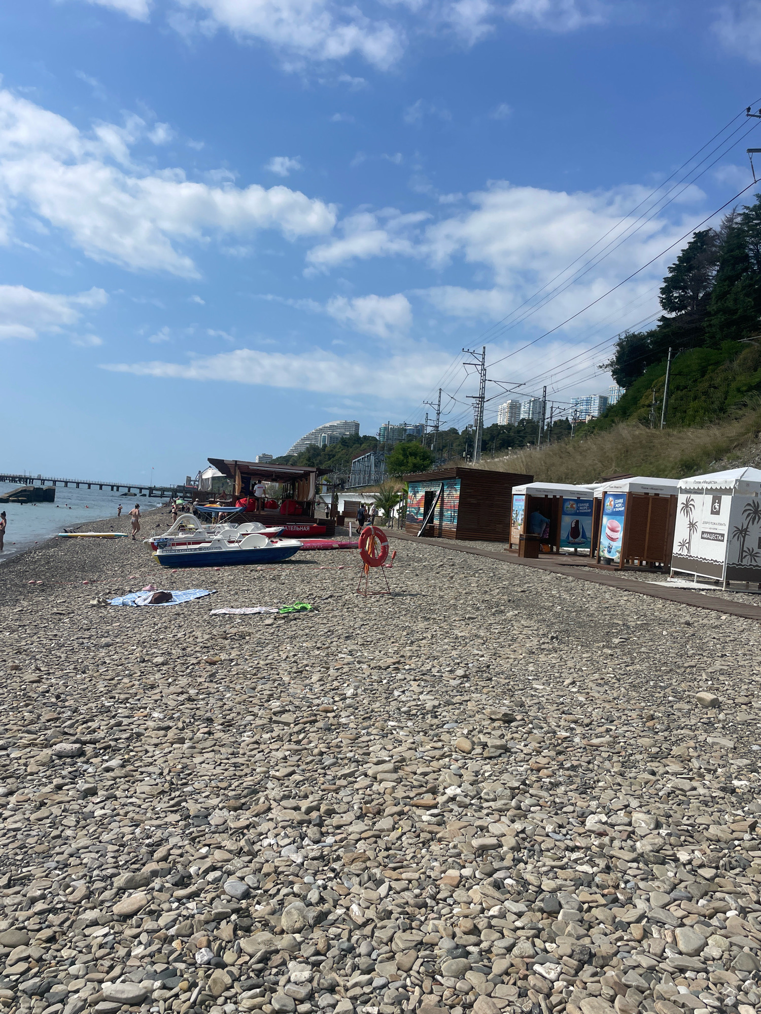 This image depicts a pebble beach in Sochi, Russia, on a sunny day with scattered clouds in a bright blue sky. Several buildings, possibly changing rooms or small shops, are set up along the beach. In the foreground, a blue and white pedal boat rests on the pebbles, along with a red life preserver on a stand. A few people are visible on the beach and in the water, some walking and others swimming or sunbathing. A pier extends into the sea in the background, with several buildings and a lush green hillside rising behind the beach. Power lines and poles are also visible against the sky, indicating the proximity of infrastructure. The overall scene suggests a typical day at a Russian Black Sea resort.