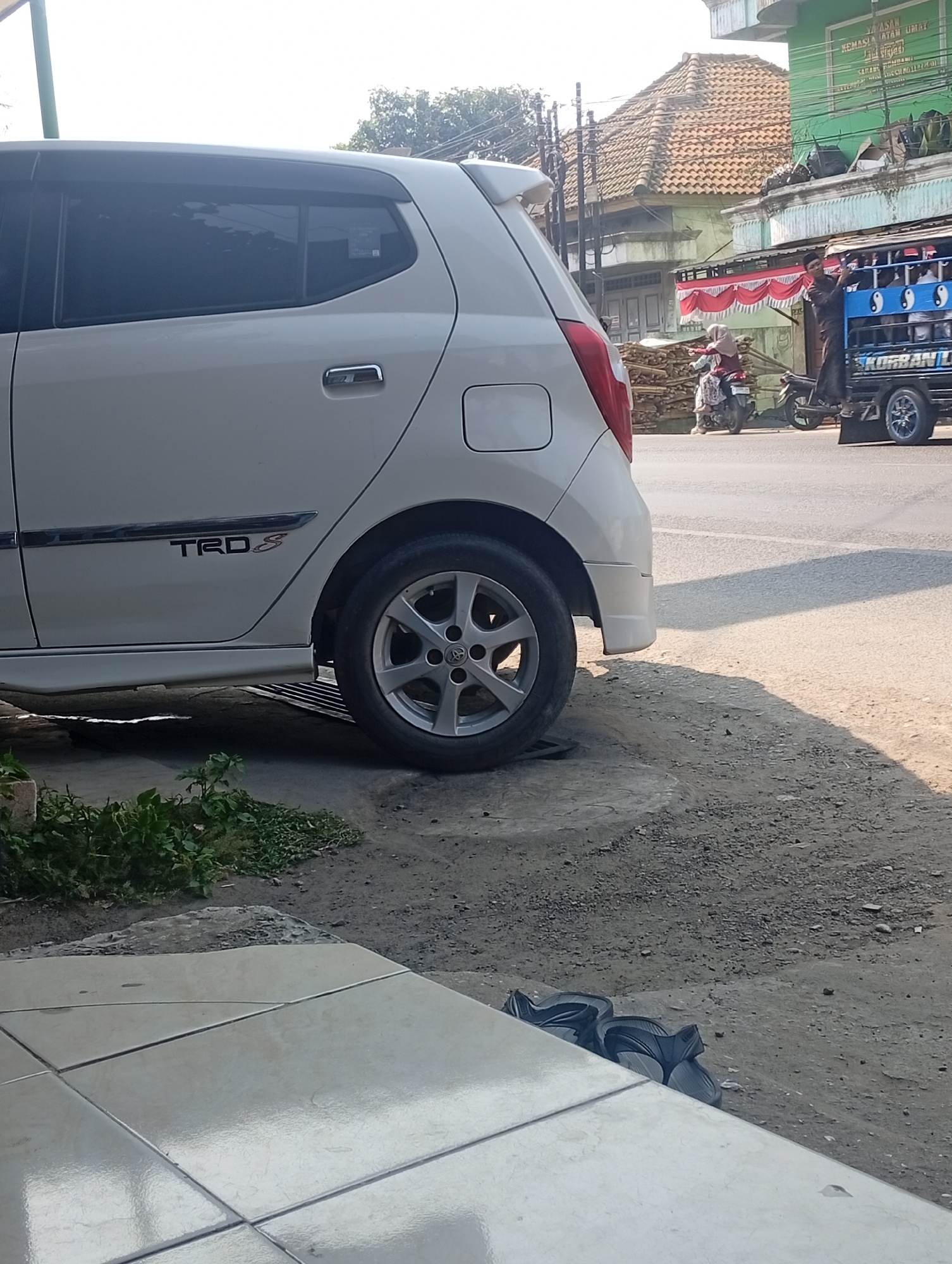 A white car with a TRD S decal is parked on a dirt shoulder next to a paved road. The car's rear passenger-side door and wheel are visible. In the background, a blue truck with people standing in the back is on the road. Adjacent to the road is a building with a green facade and a tiled roof, with red and white decorations. To the left of the car, a small patch of green plants is growing. Further in the background, a motorcycle with two riders is visible. The scene appears to be a street in Bajingjowo, Indonesia, with a mixture of residential and commercial buildings. The lighting suggests it is daytime, possibly late afternoon due to the long shadows.