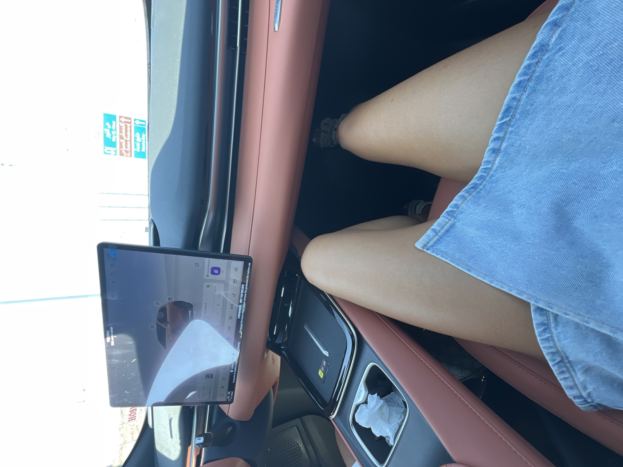 The image shows the interior of a car. The dashboard features a large touchscreen display showing a car model. A person's legs are visible in the foreground, wearing a denim skirt. In the background, through the windshield, signs for locations in Sharm El Sheikh, Egypt, are visible, including "Hey El-Nour," "Al-Salam Boulevard," and "Naama Bay." The car's interior has a pink-toned upholstery. A wireless charging pad and a cup holder with crumpled paper are also visible on the center console.