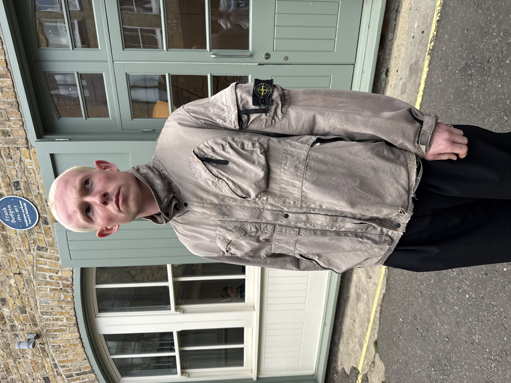 A young man with short, bleached blonde hair and light-colored eyes stands outdoors, posing for a photograph. He has a neutral expression and looks directly at the camera. He is wearing a light brownish-grey or taupe utility-style jacket with multiple pockets, including flap pockets on the chest and lower body, and a zipped pocket on the upper left sleeve. A prominent Stone Island badge, featuring a compass rose design in yellow and black, is visible on the upper left arm of the jacket. Underneath the jacket, black clothing, likely trousers, can be seen.
The background features brick buildings, typical of London architecture, with large, multi-paned wooden doors and windows painted a muted sage green. To the left of the man, a blue circular plaque is mounted on the brick wall. The text on the plaque reads "Frank Budgen," "1954-2015," and identifies him as a "Photographer and director." The ground is an asphalt surface with a yellow painted line, suggesting a street or paved walkway. The natural light indicates it is daytime, likely an overcast or mild day given the soft lighting and lack of harsh shadows. The man has a small earring in his left ear and a ring on his right ring finger.