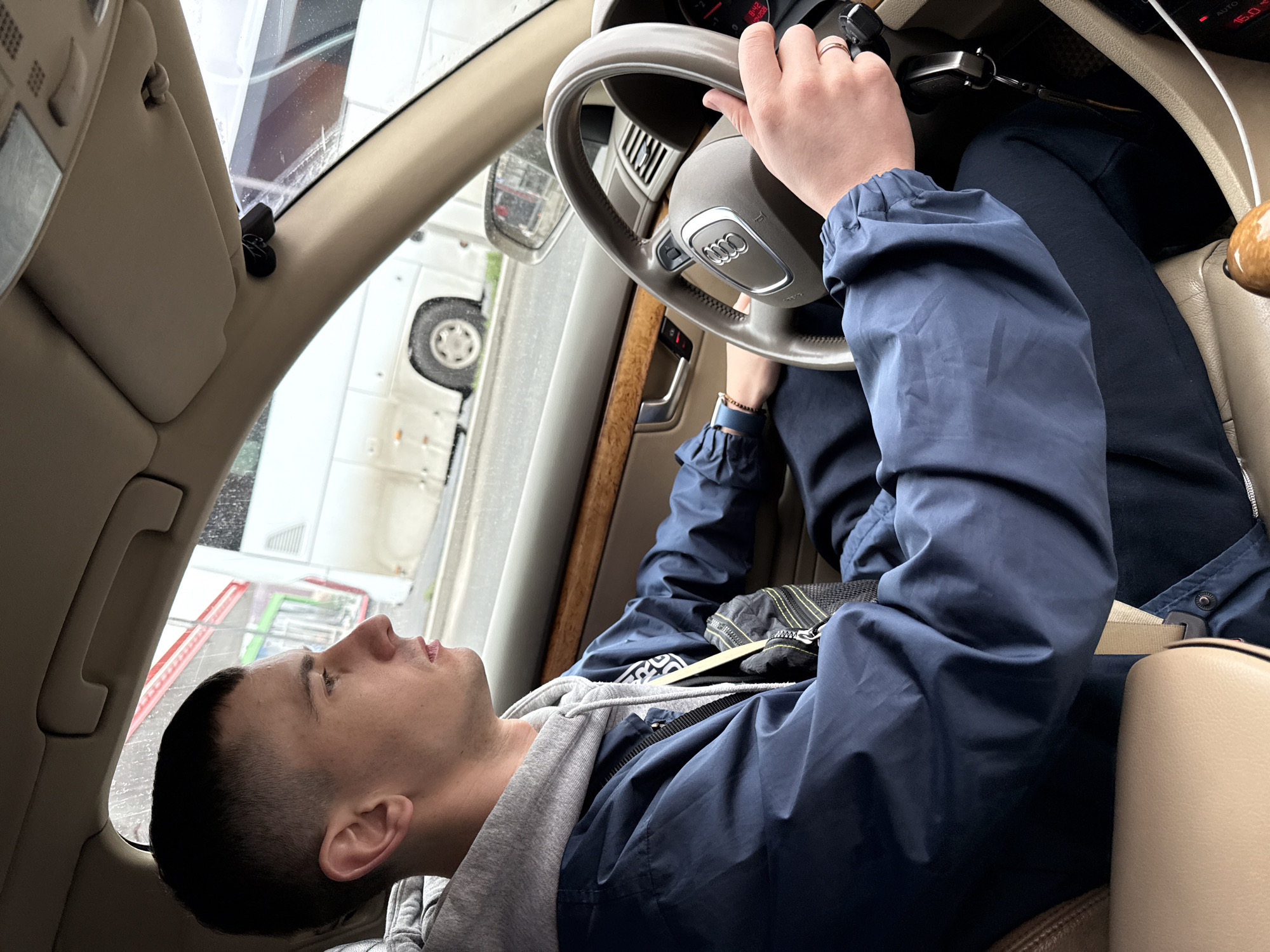 A man is lying in the driver's seat of a car, holding the steering wheel. He is wearing a blue jacket over a gray hoodie. The car appears to be an Audi. The interior of the car is beige. The car is parked outside; through the window, a glimpse of a building is visible, showing colorful stripes and a white vehicle. The setting and architecture do not definitively indicate Moscow, Russia.