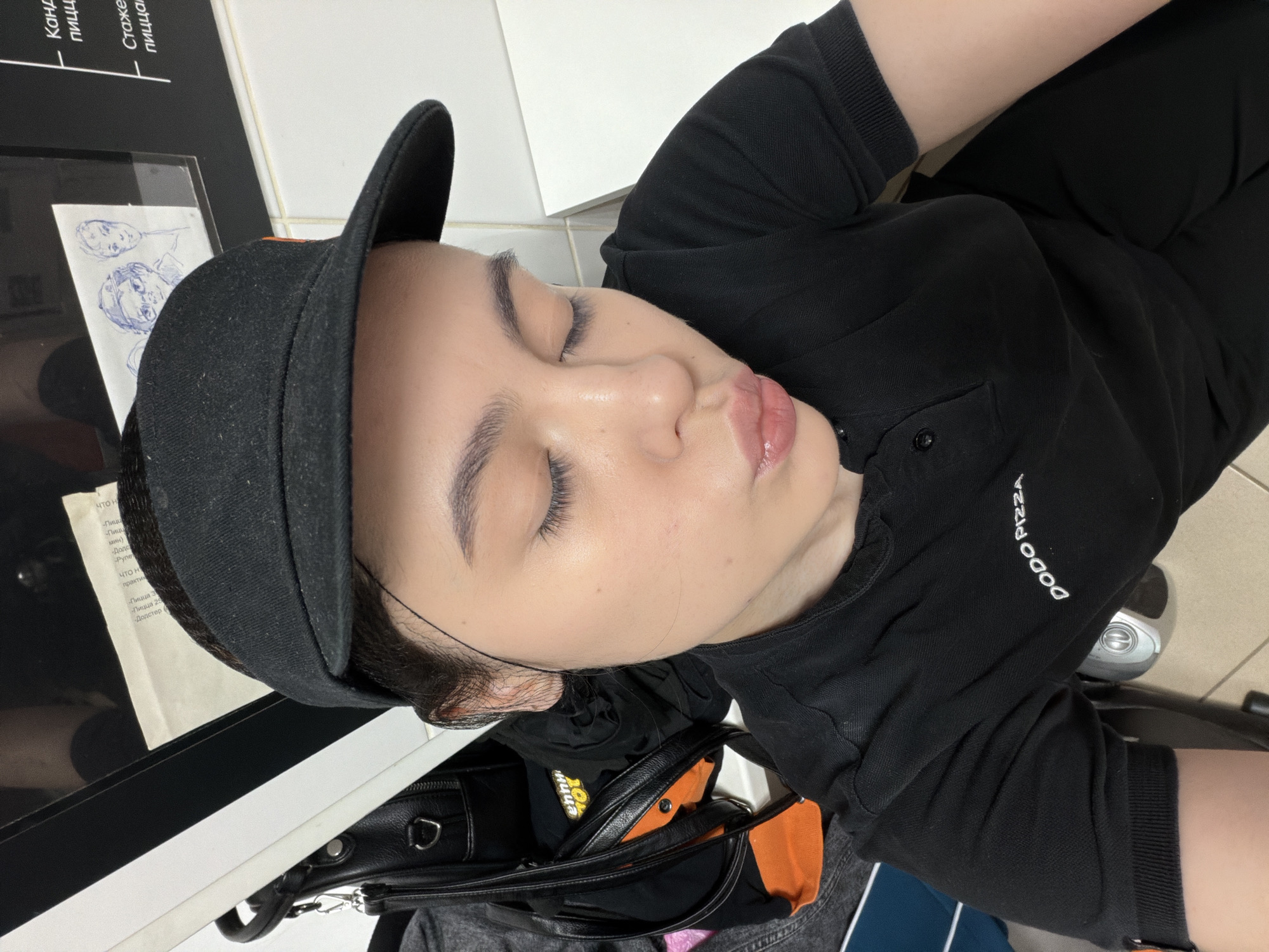 This image, taken in what is contextually identified as Chaikovsky, Russia, features a person, likely female, resting indoors. The person's eyes are closed, suggesting they are sleeping or taking a break. They are positioned leaning against a white surface, possibly a counter or wall, and are wearing a black baseball cap and a black short-sleeved polo shirt. The right sleeve of the shirt clearly displays the embroidered white text "DODO PIZZA," indicating the location is likely a Dodo Pizza restaurant or an area associated with it.
The person's face is relaxed, with closed eyelids and subtly glossed lips. Dark hair is visible around the face, peeking from under the cap. To the left of the person's head, on a dark, reflective surface, are papers with Cyrillic text and some hand-drawn sketches of faces in blue ink.
Other objects in the scene include a black and orange bag or backpack, and another dark bag situated near the person's right shoulder. The visible text from the papers includes phrases in Russian such as "Канд Пицц" (possibly "Candidate Pizza"), "Стаже пицца" ("Intern Pizza"), "Что н практик" ("What is practice"), "Пицца" ("Pizza"), and "Додстер" ("Dodster," a known Dodo Pizza menu item). The overall lighting suggests an indoor setting, possibly during the day or under bright artificial lights. The atmosphere appears calm and quiet, consistent with someone taking a moment to rest.