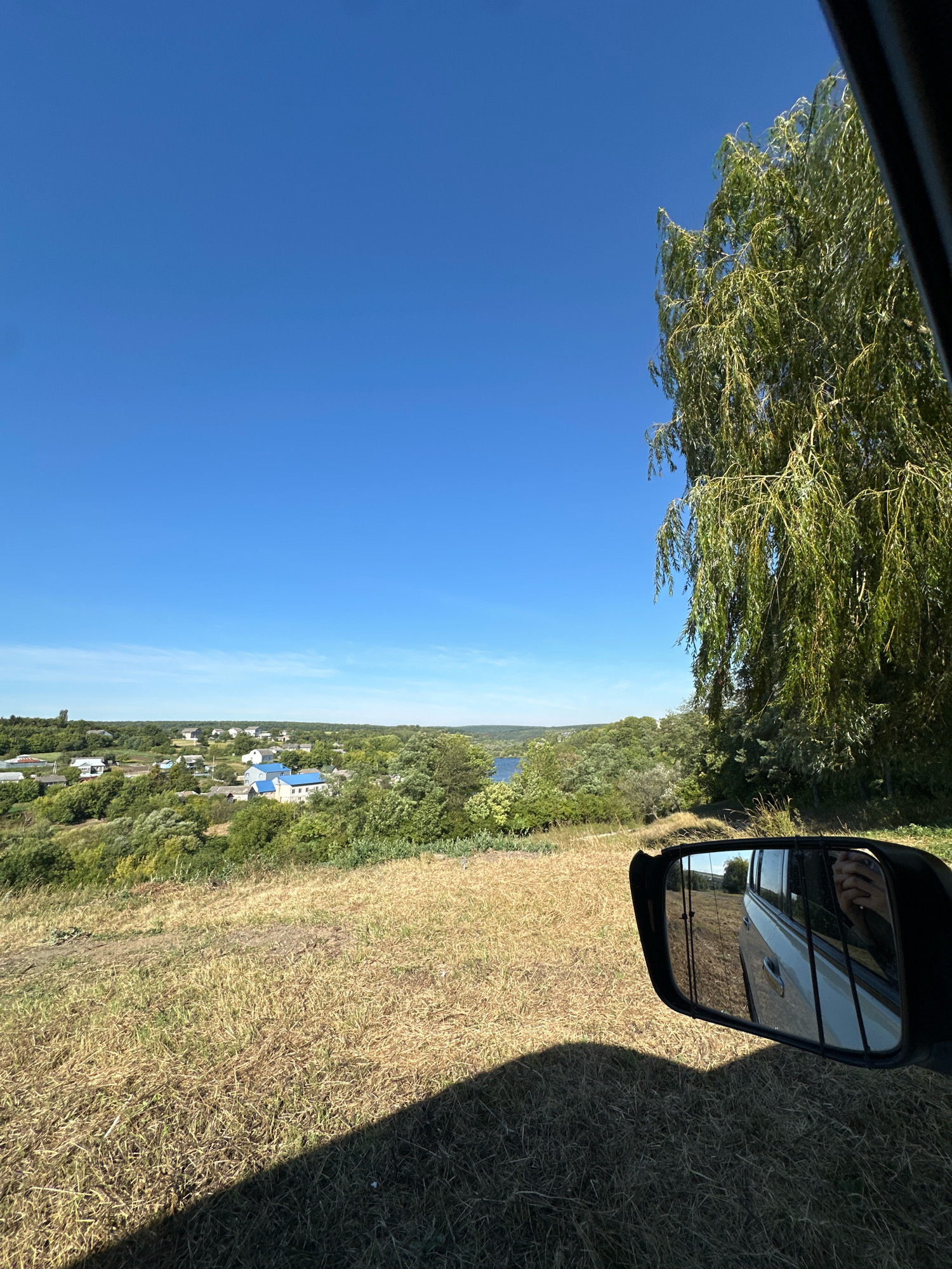 The image shows a view from inside a vehicle, looking out over a rural landscape under a clear blue sky. In the foreground, to the right, is the side mirror of the vehicle, reflecting the scene behind it, including a hand holding a phone, and the car's exterior. The ground in the foreground is covered with dry, yellowish grass. Beyond this, a gentle slope leads down to a village with scattered houses, many with blue roofs, nestled amongst trees. In the distance, a body of water, possibly a river or lake, is visible. A large tree with drooping branches is prominent on the right side of the image, obscuring part of the sky. The overall impression is of a sunny day in a countryside setting, likely in or near the village of Makiv, Ukraine, given the context.