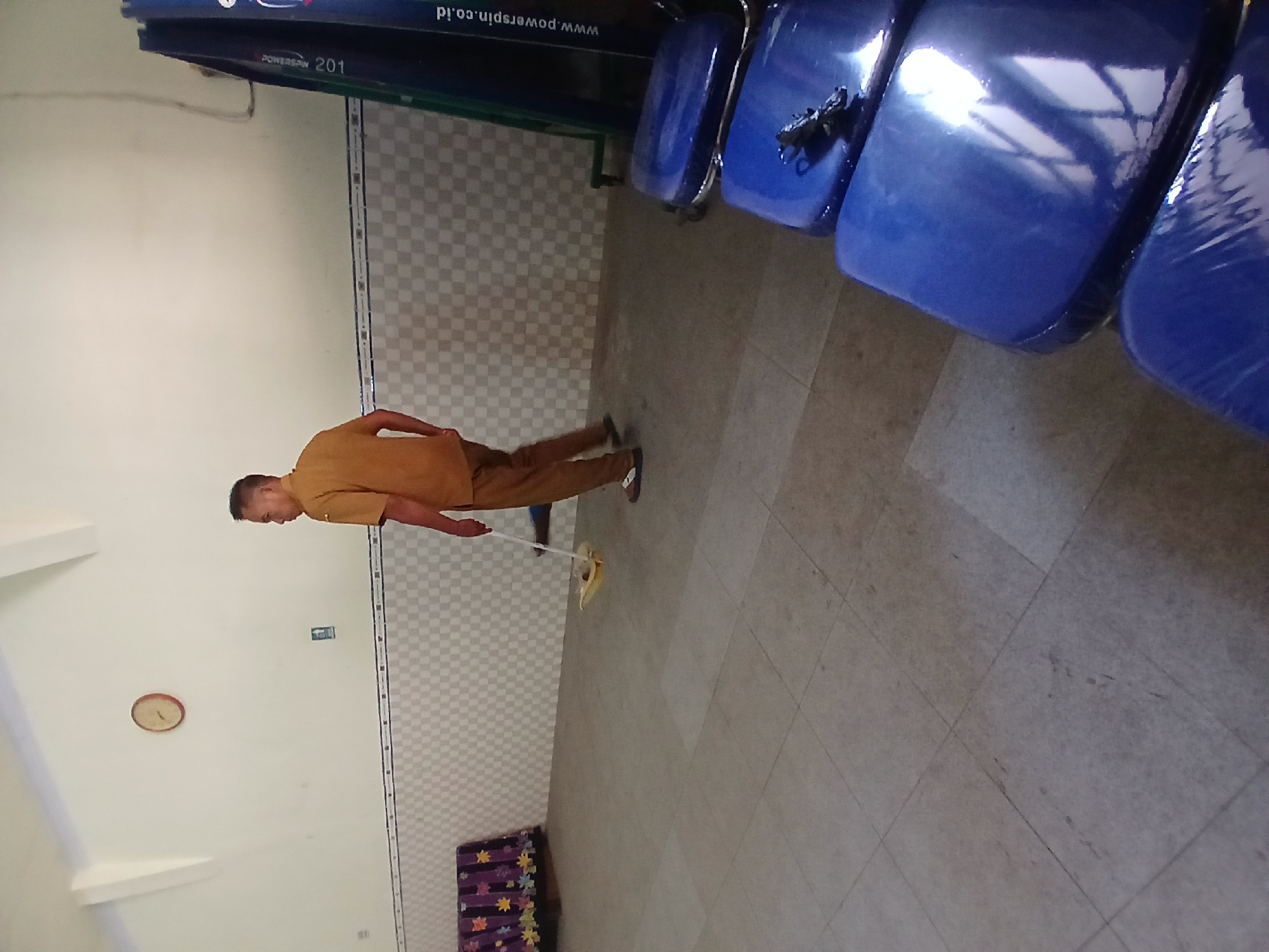 A man wearing a tan uniform and brown pants is mopping the tiled floor in a room. He is facing away from the camera and towards the left side of the image. To his left is a piece of furniture covered with a patterned cloth. In the background, a clock is mounted on the wall. To the right of the man, a dark green rack with blue seats is visible. The walls are tiled with a checkered pattern. The room appears to be indoors, possibly a community hall or sports facility in Pejaten, Indonesia.
