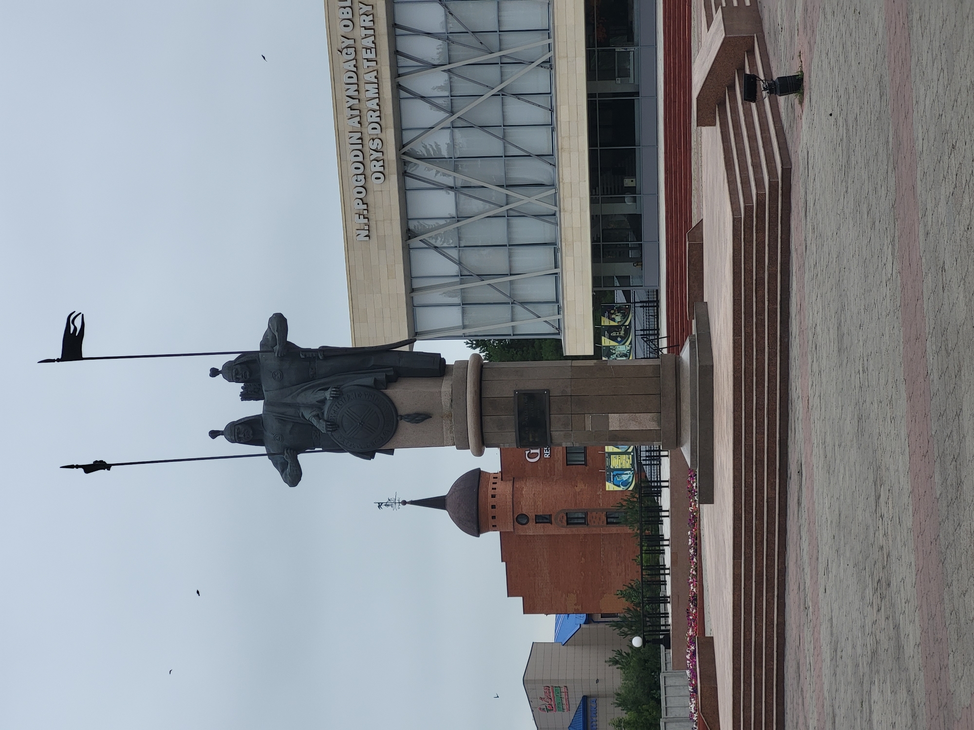 The image shows a monument in front of the N.F. Pogodin Regional Kazakh Drama Theater in Petropavl, Kazakhstan. The monument is a bronze statue of two warriors in historical armor, holding spears and a shield. They are standing on a stone pedestal, which is surrounded by a tiered platform. In the background, the theater building is visible with its modern facade, featuring large windows and a sign displaying the theater's name. To the left of the monument, there is a brick building with a domed tower. The ground in front of the monument is paved with stone tiles. The sky is overcast. There are no people clearly visible in the immediate vicinity of the monument.