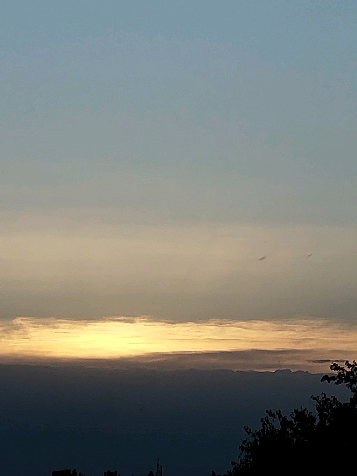 The image depicts a sky with layered clouds at sunset or sunrise. The upper portion of the sky is a clear, pale blue, gradually transitioning to a band of soft yellow and orange clouds. Below this, a darker, more diffused layer of clouds stretches across the horizon, appearing almost purple or grey. The very bottom of the image shows the silhouetted outlines of trees and what appears to be the tops of buildings or structures, suggesting an urban environment. There are no visible people or distinct objects within the clouds. The overall scene conveys a serene and atmospheric natural phenomenon. Based on the presence of what look like cell towers and building silhouettes, it is possible this image was taken in the city of Bucharest, Romania, during twilight.
