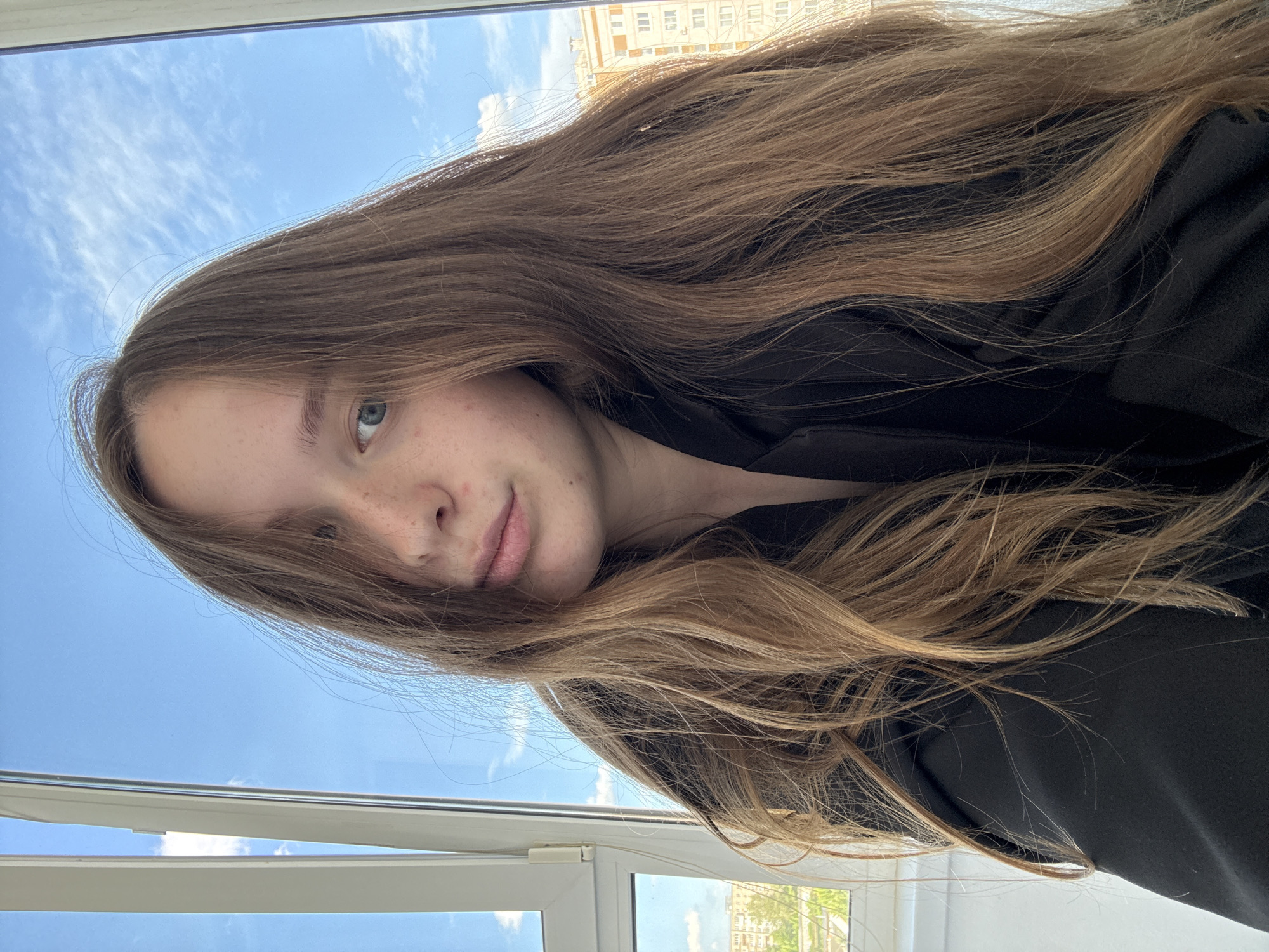 A young woman with long, light brown hair and fair skin with freckles is shown from the chest up, looking towards the camera. She is wearing a black jacket. Her hair is partially covering her face. She is positioned in front of a window, with a view of a blue sky with clouds and buildings visible outside. The window frame is white. The scene appears to be indoors, likely in an apartment or house in Chaikovsky, Russia, given the architectural style of the buildings in the background.
