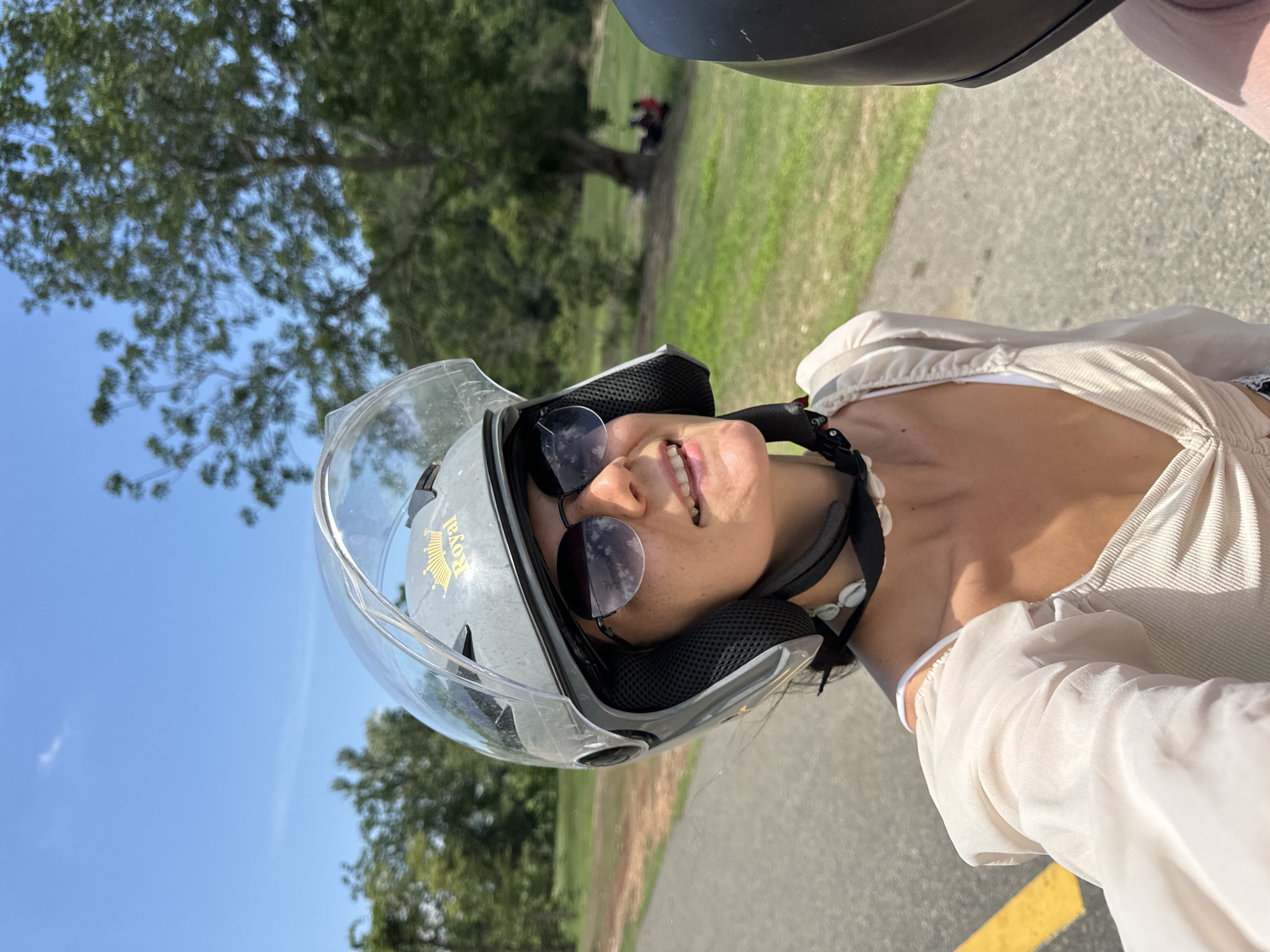 A woman wearing a grey helmet with a clear visor and "Royal" logo is riding on a paved road in Ham Thuan Bac, Vietnam. She is wearing sunglasses and a light-colored top with a seashell necklace. The road has a yellow line marking on the left side. The background consists of lush green trees and grass under a clear blue sky. In the distance, a group of people can be seen sitting on the grass near a large tree. The image is a selfie, taken from the perspective of the rider.