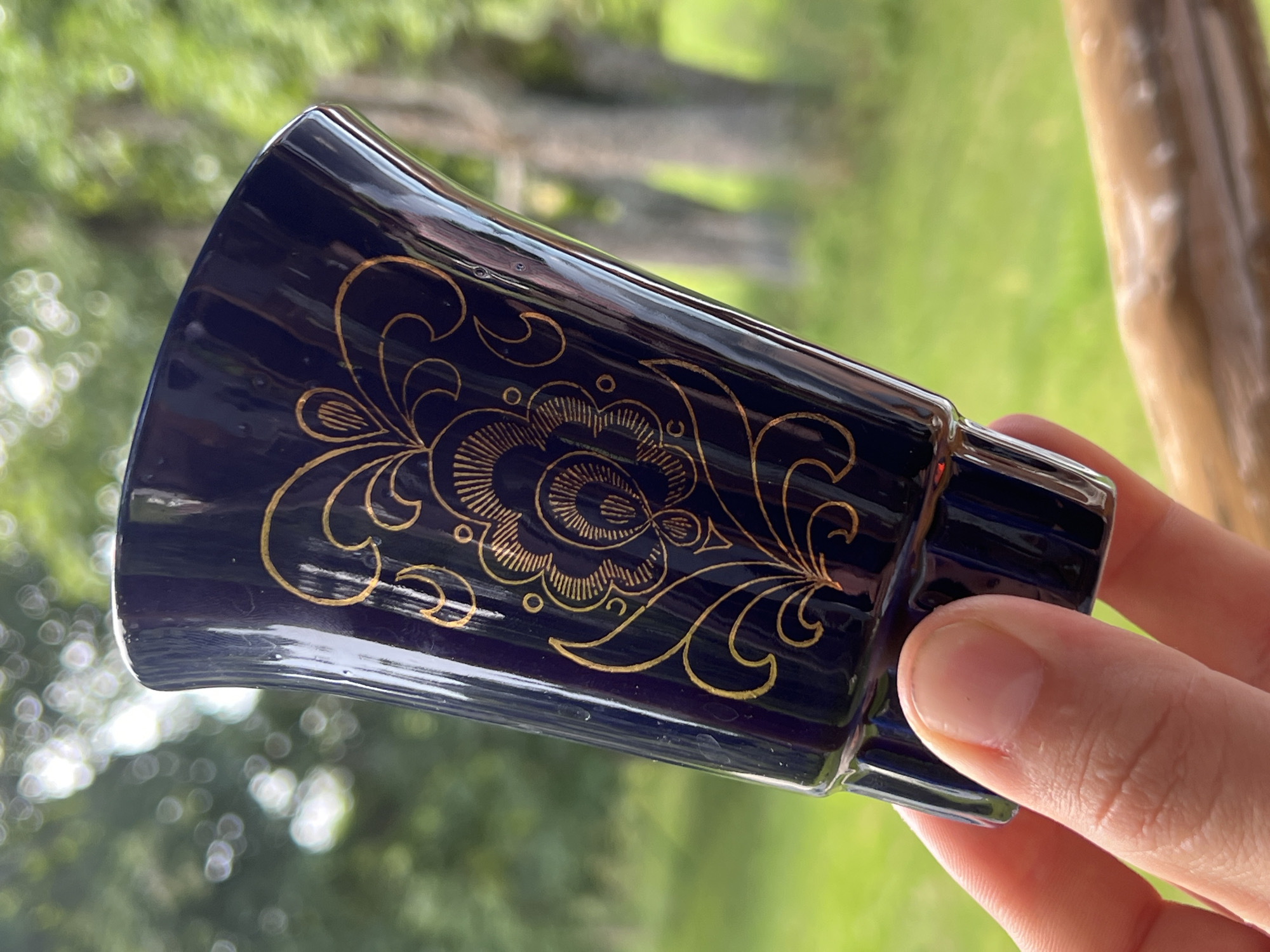 A close-up, eye-level shot shows a hand holding a dark blue, ornate ceramic cup with gold floral accents. The cup is positioned at an angle, with its flared opening facing upwards and to the right. The gold design on the cup features a central flower-like motif surrounded by swirling, vine-like patterns. The background is blurred and out of focus, showing a lush green lawn and trees with dappled sunlight, suggesting an outdoor setting. The image was taken at the Norviliškės Castle complex in Lithuania.