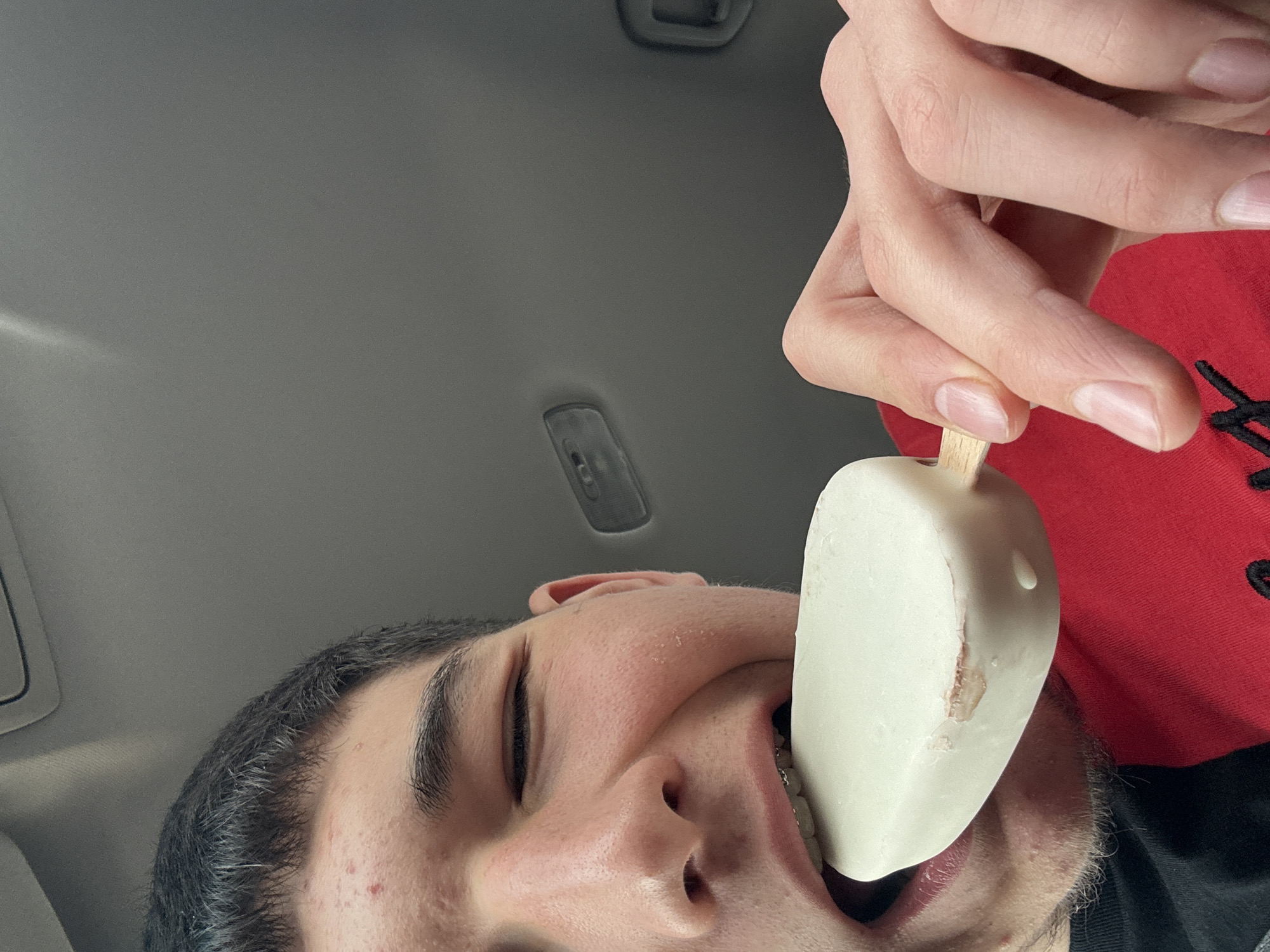 An upward-facing close-up shot captures a person eating an ice cream bar inside what appears to be a vehicle in Dobromirtsi, Bulgaria. The person's face is visible from the nose down, angled from the bottom left of the frame, with the top of their head and a gray car ceiling in the upper left. Their eye is closed or heavily squinting, and their mouth is open, revealing braces on their lower teeth, as they are about to bite into or are already biting a white, rectangular ice cream bar.
The ice cream, held by a wooden stick gripped by the person's right hand from the top right of the frame, is coated in white and shows a slight blemish or melt mark on one side. The person has dark hair, stubble around their chin, and some minor redness on their cheek. They are wearing a red garment, possibly a shirt or hoodie, visible on the right side. The vehicle's interior features a light gray ceiling with a dark fixture (possibly a handle or light) visible towards the top. There is no visible text in the image.
