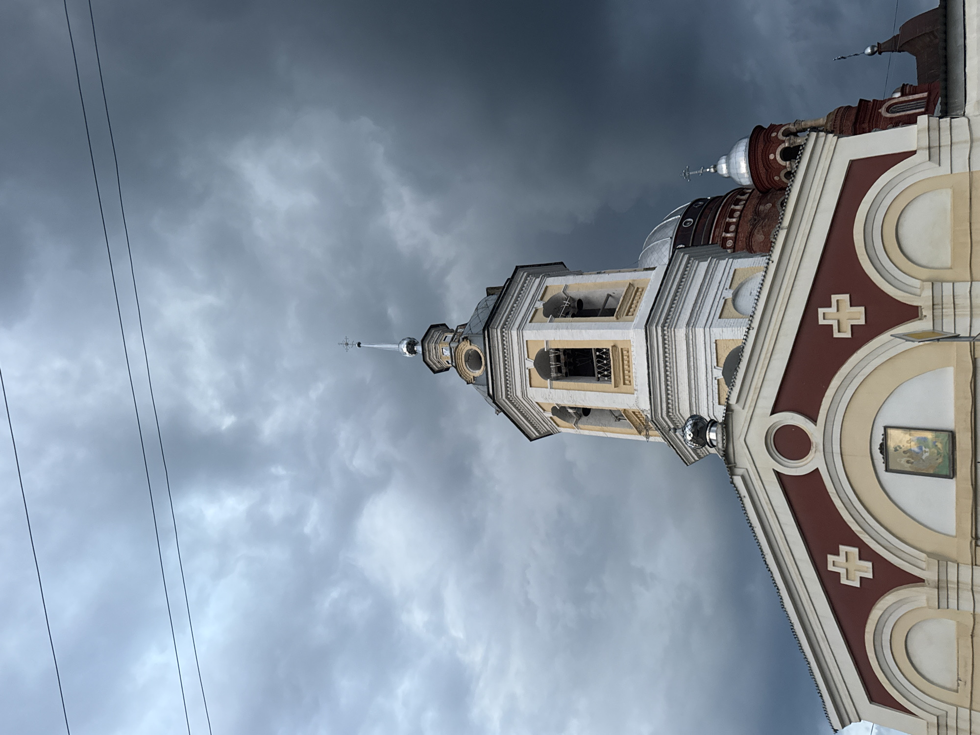 An upward angled, medium shot shows the top of a Russian Orthodox church building against a dramatic, dark, cloudy sky. The architecture features a white and yellow bell tower with arched openings and bells visible within. Below the bell tower, part of the main church building is visible with a red and white facade decorated with crosses and a religious icon. The sky is filled with heavy, gray clouds, suggesting an approaching storm. A few thin electrical wires are strung across the upper portion of the frame. No people are visible in the image. The overall impression is one of solemnity and impending weather. The building is the Transfiguration Cathedral in Urzhum, Russia.