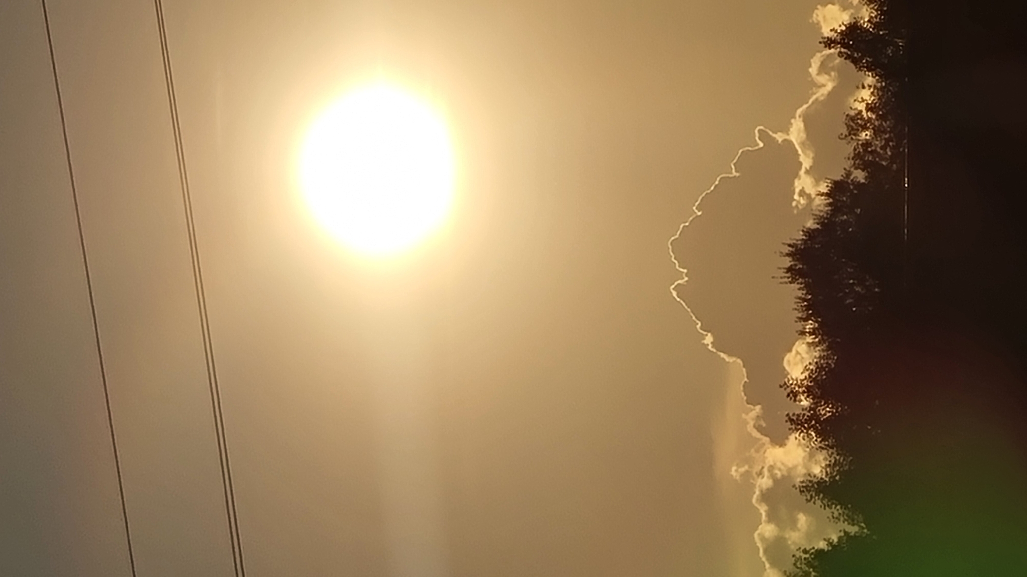 The image captures a sunset over Vysokoe, Russia, with the sun appearing as a bright, luminous orb near the top center of the frame. Two parallel power lines stretch horizontally across the upper portion of the sky. Below the sun, dramatic clouds are illuminated by the fading light, with one large cloud in the center having a distinct, jagged outline rimmed with light. A dense line of trees forms a dark silhouette along the bottom of the image, obscuring the ground and any potential urban features. The overall atmosphere is subdued and serene, dominated by the warm hues of the sunset.