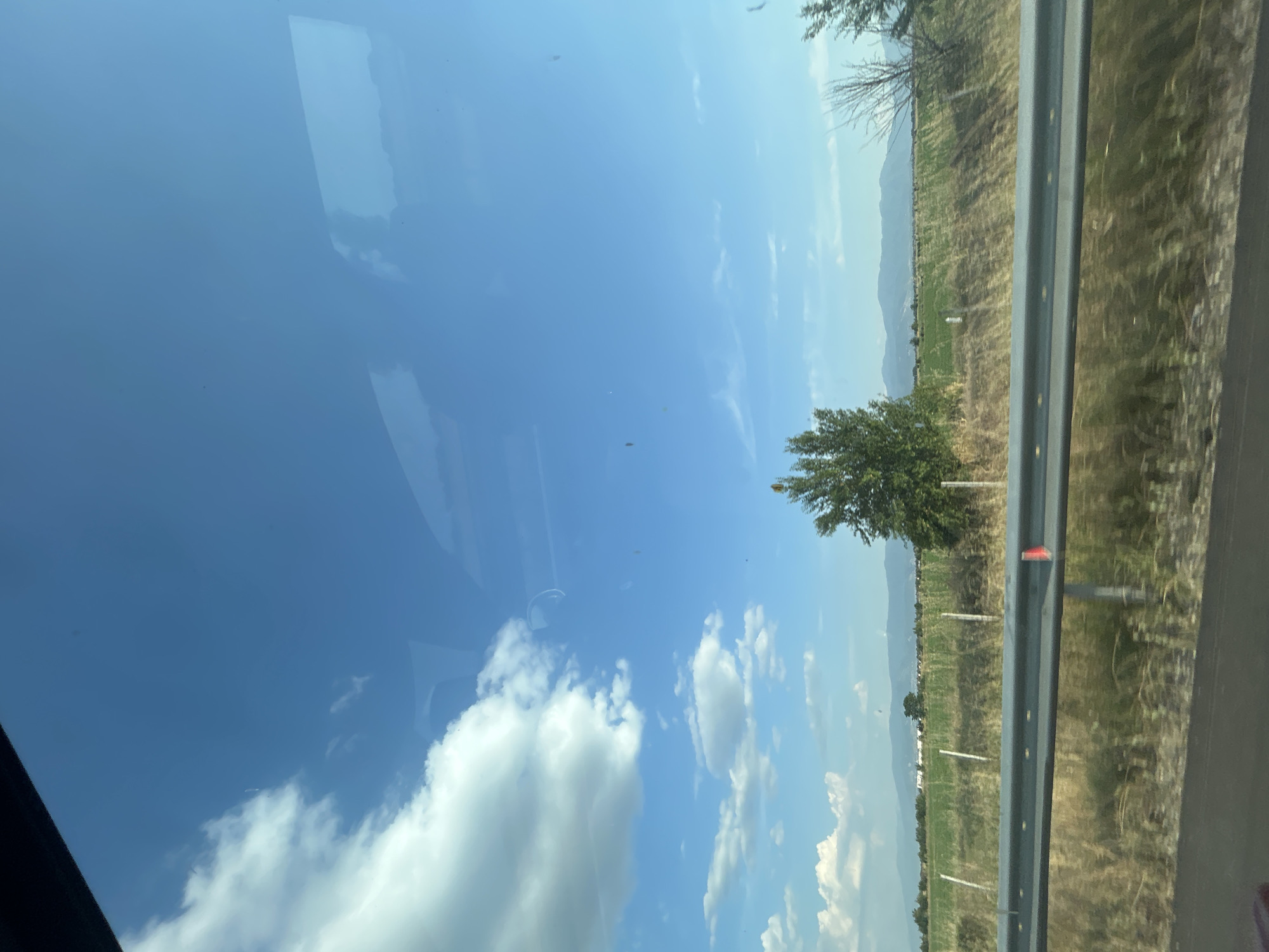 The image depicts a landscape scene, likely taken from inside a vehicle, as suggested by the reflection on the glass in the foreground. The perspective is slightly tilted. The dominant elements are a vast blue sky with scattered white clouds and a distant landscape.
In the mid-ground, there is a line of trees and a green field, with a prominent single tree standing out. Beyond the field, a mountain range is visible, fading into the haze. A grey metal guardrail runs along the right side of the image, indicating a road or highway. The vegetation on the right side of the guardrail appears dry and brownish. The scene suggests a rural or semi-rural setting.
The lighting indicates it is daytime, likely late morning or early afternoon, given the bright sun and clear blue sky, though some clouds are present. The weather appears to be pleasant and sunny. There are no people or explicit man-made structures other than the guardrail and what appears to be a distant fence line. The overall mood is tranquil and expansive, conveying a sense of travel or observation of the passing scenery.
No visible text is present in the image. The location context provided places this scene near Plovdiv, Bulgaria.