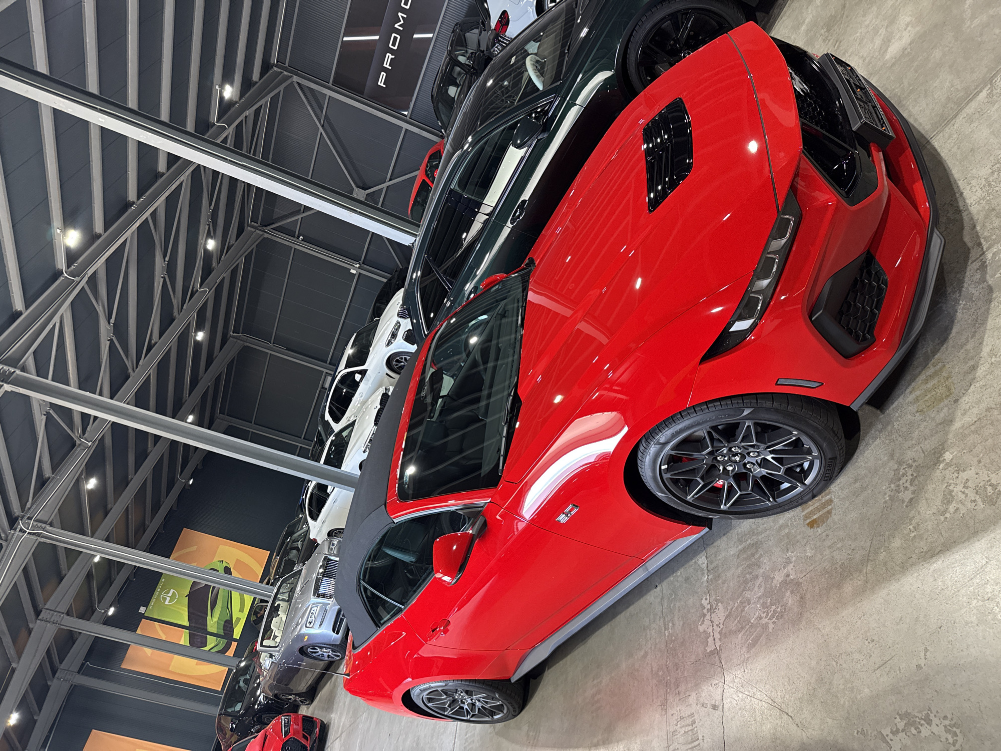 The image shows a red convertible sports car parked indoors, possibly in a car dealership, with other vehicles visible in the background. The car is facing towards the right of the frame and occupies the foreground. It has a black convertible top and black accents on its exterior, including the hood and wheels. The wheels are large and have a multi-spoke design. The car's front end is visible, featuring sharp headlights and a prominent grille. The environment appears to be a modern building with a high ceiling supported by metal beams. The floor is concrete. In the background, several other cars are parked, including a silver convertible, a dark-colored SUV, and white SUVs. There are also large banners with graphics of cars and text hanging on the back wall. The scene is well-lit, suggesting an indoor showroom. The location is stated to be in Plovdiv, Bulgaria. There are no people visible in the image.