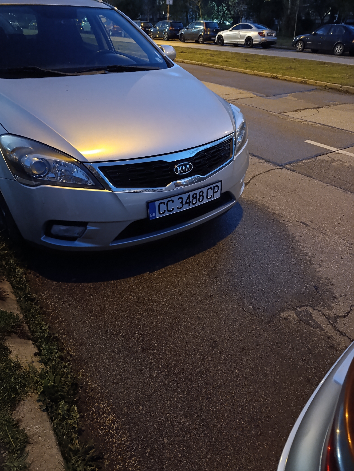 The image shows a silver Kia Cee'd parked on the side of a street. The license plate reads "CC 3488 CP" with the country code "BG" indicating it is registered in Bulgaria. The location appears to be urban, likely a residential area, evidenced by the sidewalk, grass verge, and other parked vehicles in the background. Judging from the license plate format and the overall environment, this image is likely located in Sofia, Bulgaria. The lighting suggests it is either dusk or dawn.