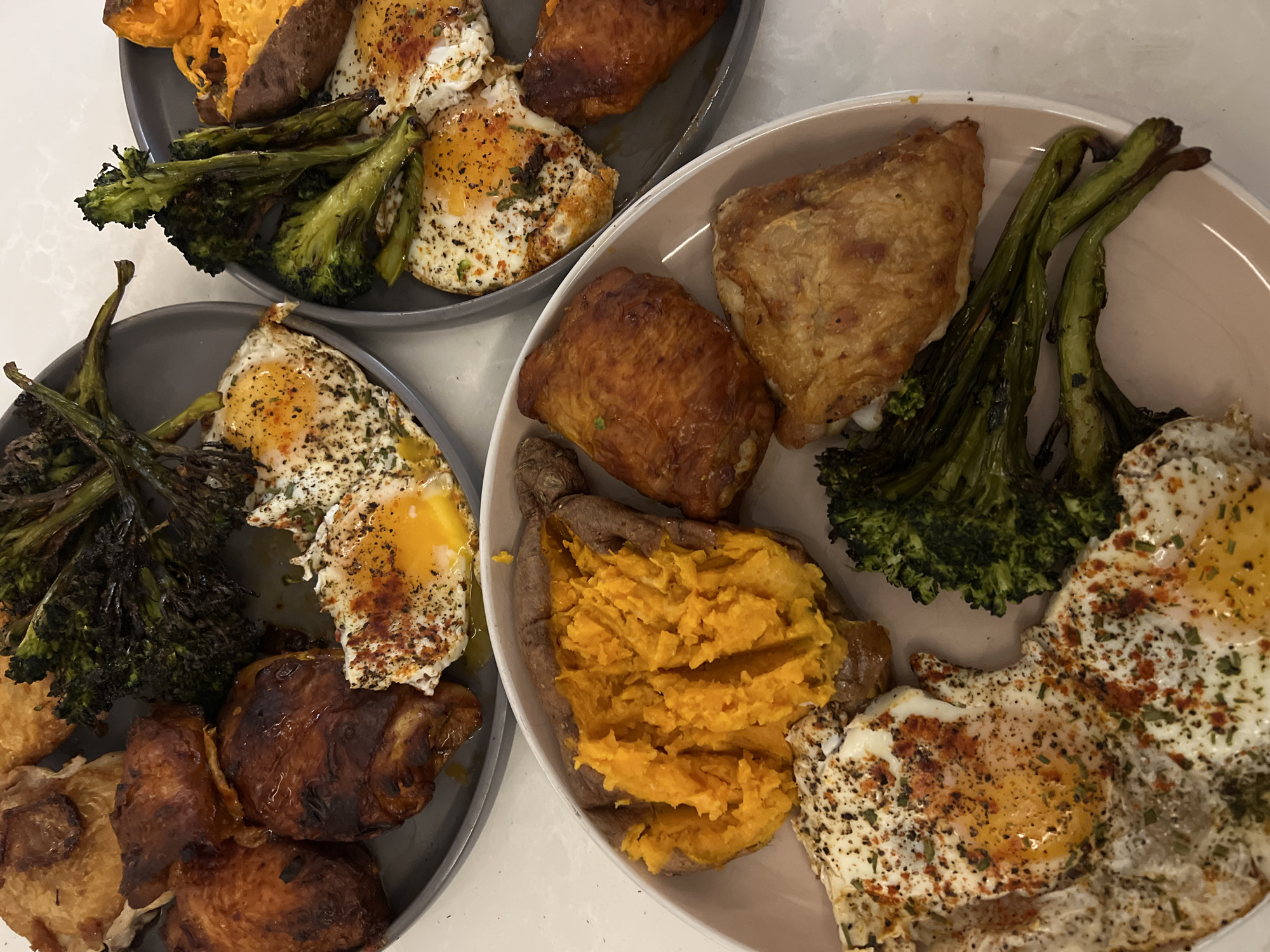 The image shows two plates of food, each containing fried eggs, roasted broccoli, baked sweet potato, and what appears to be roasted chicken pieces. One plate is gray, the other a light tan color. The food is arranged in a somewhat haphazard manner. The background is a plain, light-colored surface. There is no evidence in the image to suggest any location in London, United Kingdom or any people.