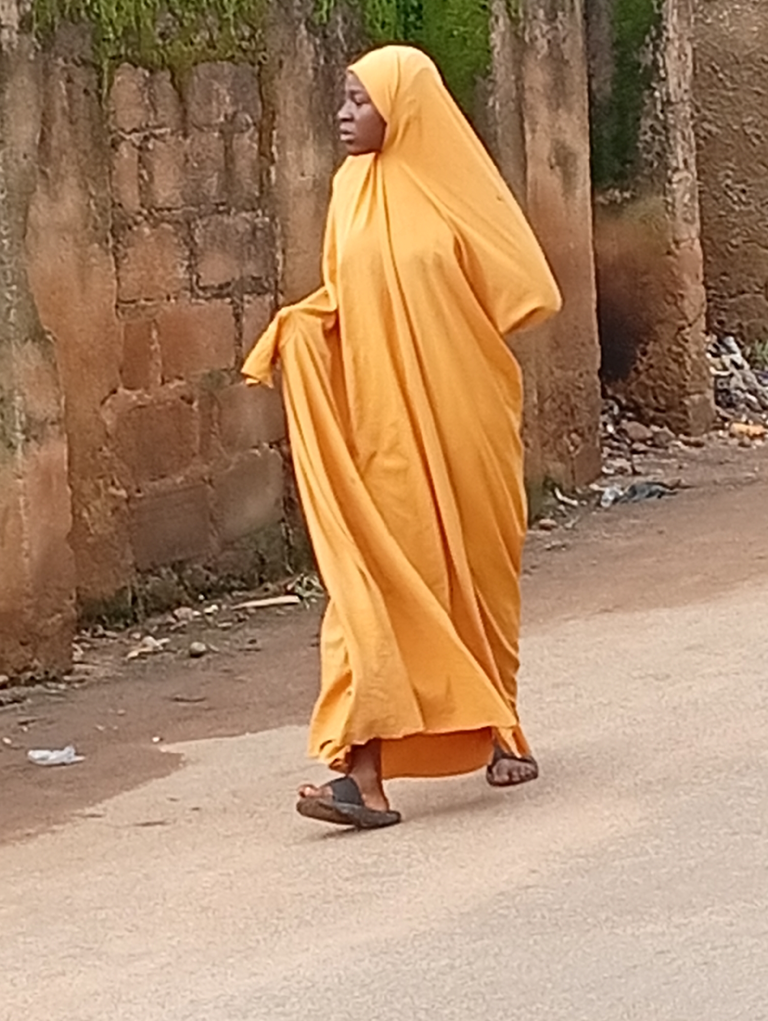 A person is walking on a dirt road in an outdoor environment. The person is wearing a bright orange headscarf and a flowing orange garment. They are also wearing dark open-toed sandals. The background consists of a rough, light-brown textured wall made of bricks and possibly concrete, with some greenery growing on top. The ground in front of the wall is a mixture of dirt and small stones, with some debris scattered around. The road is made of a light-colored, textured surface. The lighting suggests it is daytime. The scene is likely located in Jos, Nigeria, given the typical architecture and environment found in the region.