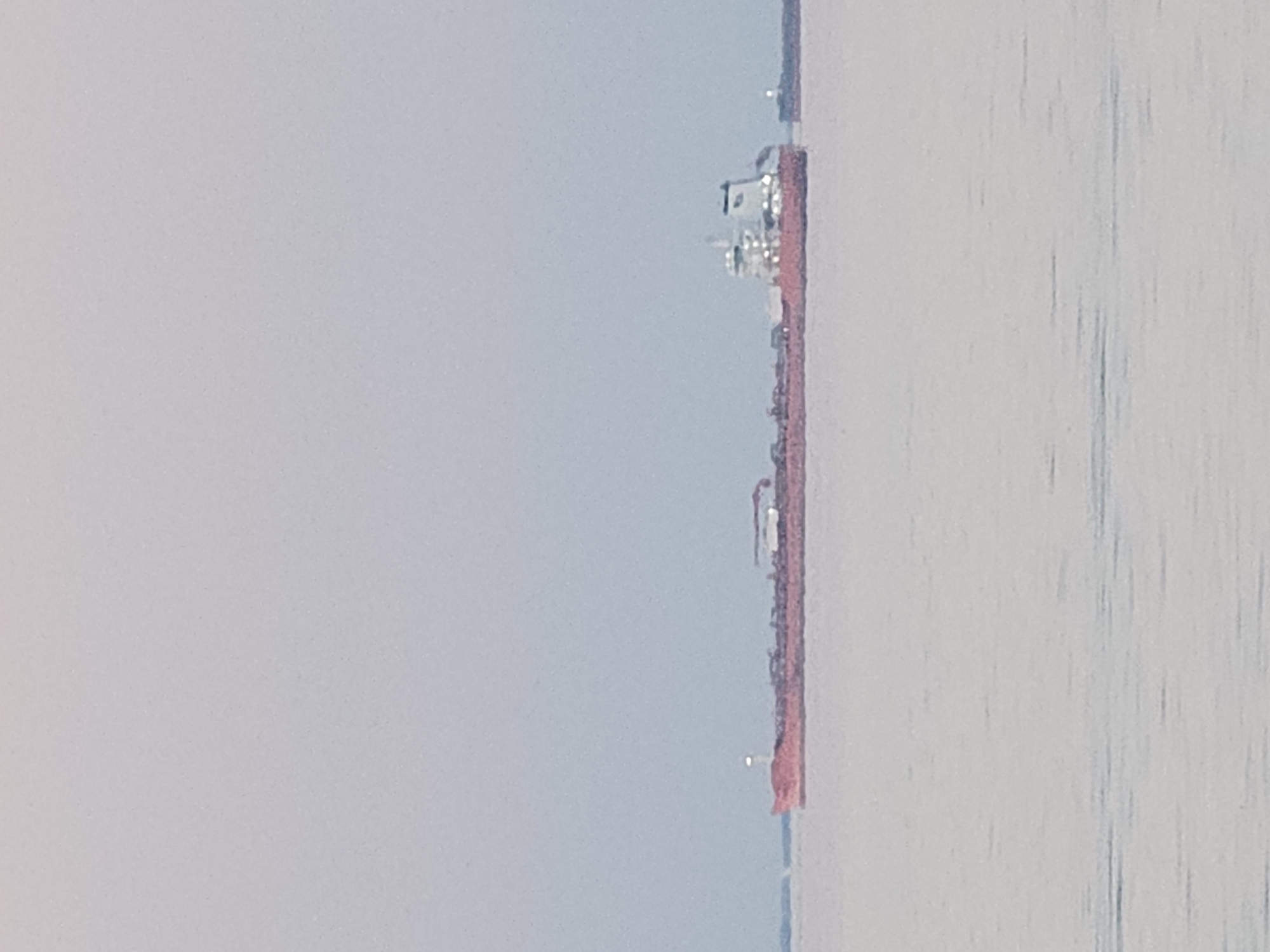 The image shows the bridge of Île de Ré, located near La Brée-les-Bains, France. The bridge appears as a thin red line in the distant view. The sky is overcast. The water of the Pertuis Breton strait is visible on the right side of the image. The view is slightly angled.