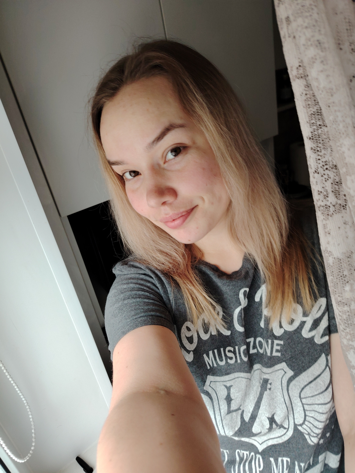 A forensic description of the scene, located in a room in the city of Марковский, Russia, reveals a young adult female captured in a close-up, chest-up selfie. The subject has light-toned skin, long, straight blonde hair with visible darker roots or undersides, and brown eyes. She exhibits a slight, closed-mouth smile. She is dressed in a grey short-sleeved t-shirt featuring a white graphic print that includes the text "ROCK & ROLL" and "MUSIC ZONE," centered above a shield emblem flanked by wings, which incorporates a guitar silhouette. Partially visible text below reads "STOP ME N". Her right arm is extended forward, holding the camera. The indoor environment includes a white vertical element on the left, consistent with a window or door frame, beside which a vertical string of white beads is present. On the right, a light-colored, patterned, translucent curtain is partially visible. The background immediately behind the subject is blurred and consists of varying shades of grey and dark tones, suggesting indistinct interior elements or furniture.