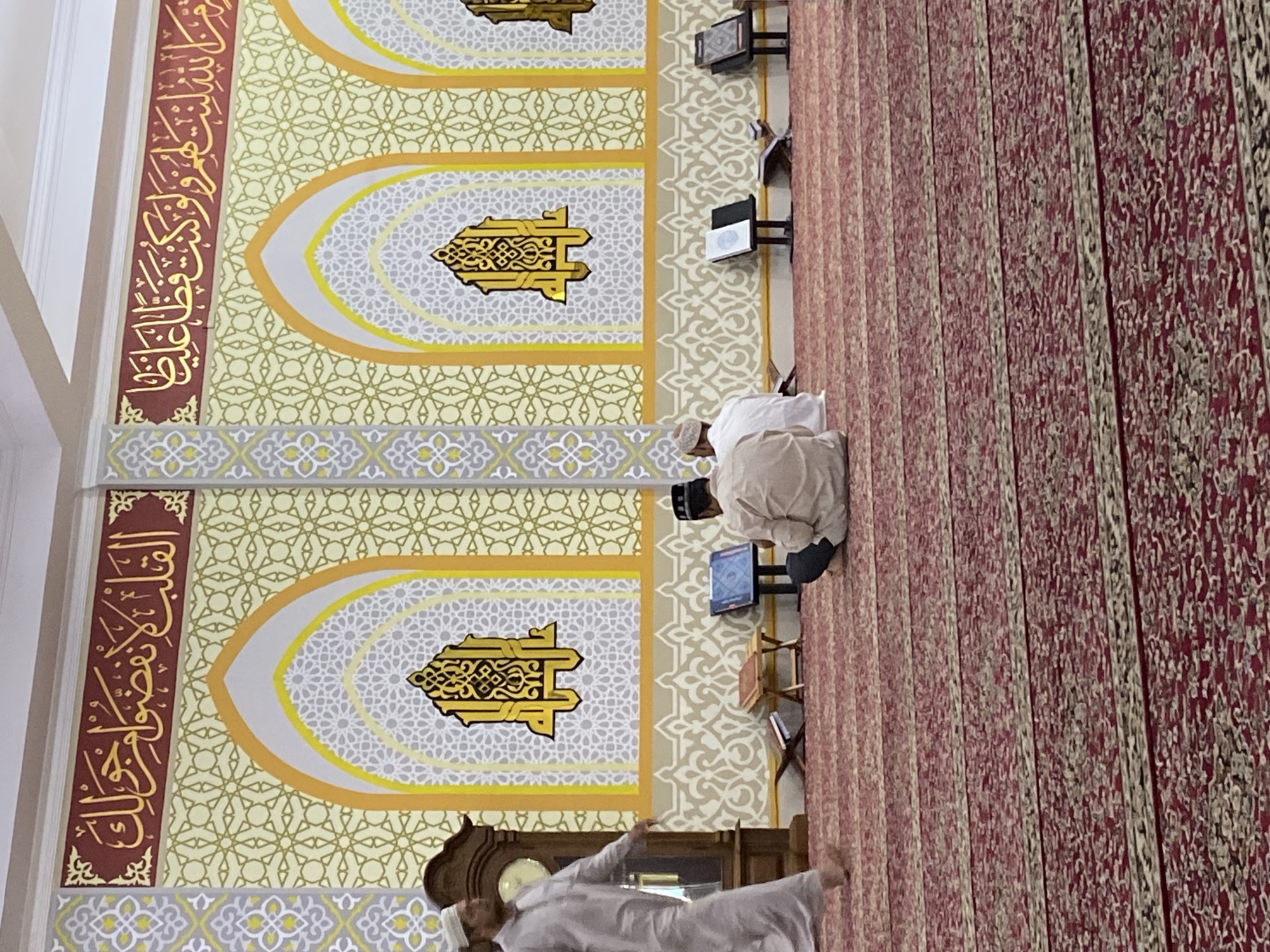 This image depicts the interior of a mosque in Lhokseumawe, Indonesia, characterized by its ornate Islamic calligraphy and architectural designs. The walls are adorned with intricate, repeating geometric patterns in yellow and white, overlaying a cream-colored background. Above these patterns, a frieze of Arabic calligraphy in gold lettering is prominently displayed, framed by a deep red border. Three arched niches, each containing a stylized golden emblem, are visible along the wall.
On the floor, which is covered with a red carpet featuring a repeating abstract pattern, several individuals are present. In the foreground, two men are seated on the carpet, facing away from the camera, seemingly engaged in prayer or contemplation. A third man, wearing a white robe and cap, walks from left to right across the scene. To the left, a tall, grandfather clock stands near the walking man. In front of the seated individuals are several low wooden stands holding books, suggesting a space for religious study or reading. The lighting appears to be artificial, illuminating the interior of the mosque. The overall impression is one of serene religious devotion within a richly decorated space.