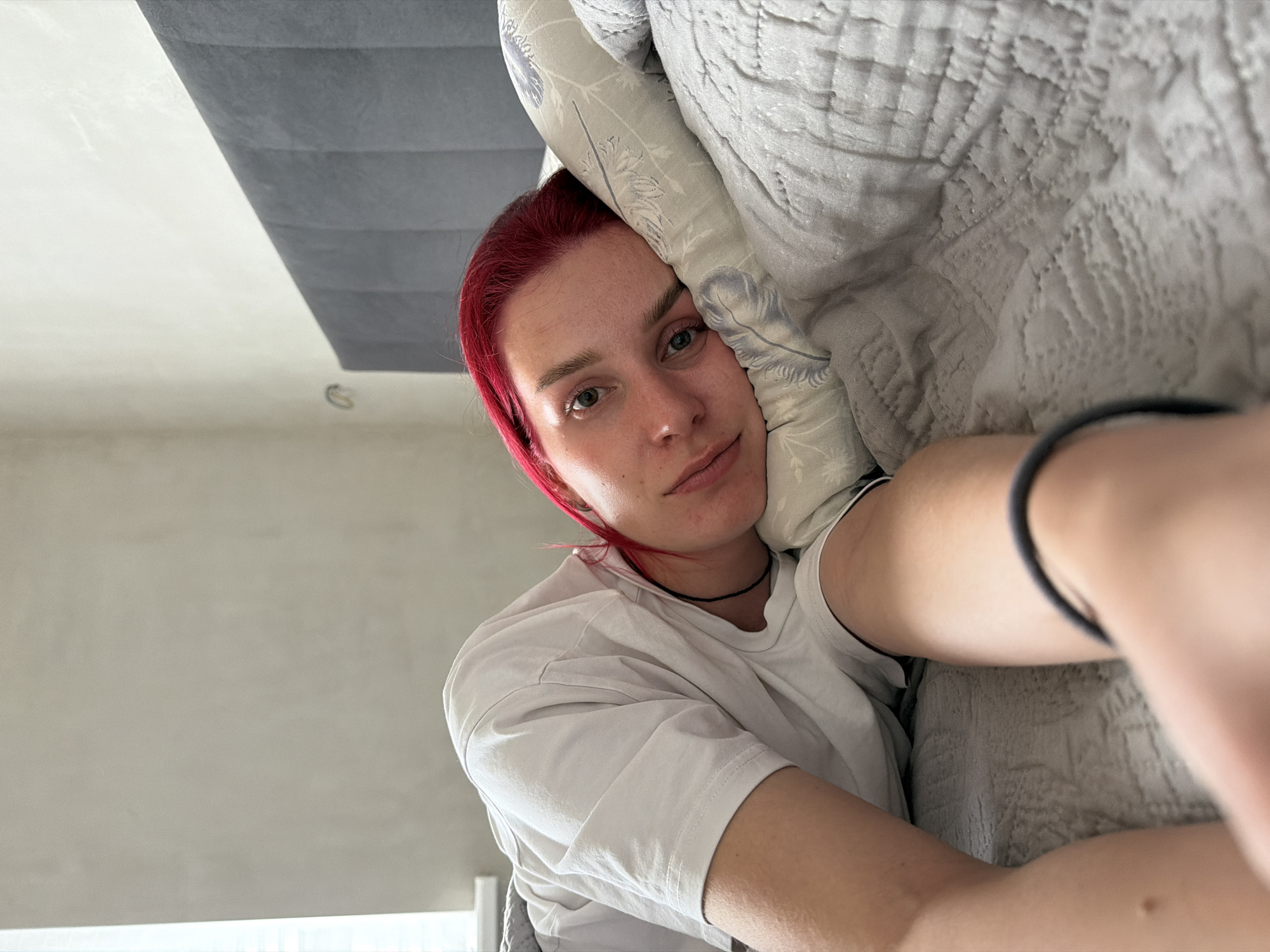 A person with red hair rests their head on a pillow in an interior location. The person is wearing a white t-shirt and a black choker necklace. The background includes a ceiling and a wall painted in neutral tones. The specific location in the city of Chaikovsky, Russia, is not discernible from the image.