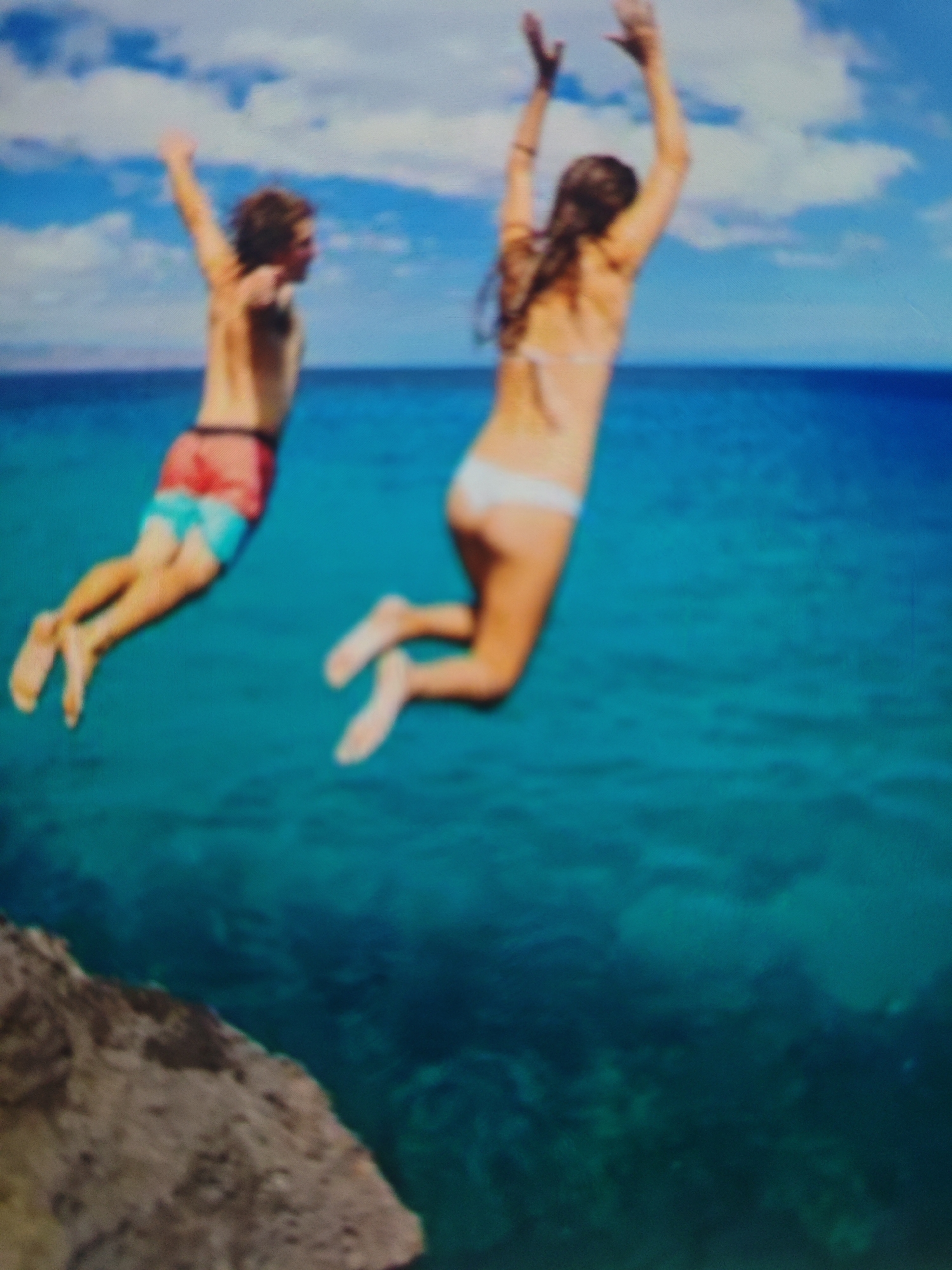 The image depicts two individuals in mid-air, jumping into a body of clear, blue-green water. The person on the left is a male, wearing red and blue patterned swim trunks. He is positioned slightly lower and to the left of the female. The female on the right is wearing a white bikini. Both are captured in motion, with their arms raised. The scene appears to be a natural environment with a rocky outcrop in the foreground on the left, suggesting a cliff or elevated diving spot. The water is a vibrant turquoise, indicative of shallow, clean tropical or subtropical waters. The sky above is blue with scattered white clouds. The overall impression is one of exhilaration and freedom, typical of summer recreation. The image does not depict any elements related to the city of Krasnaya Polyana, Russia.