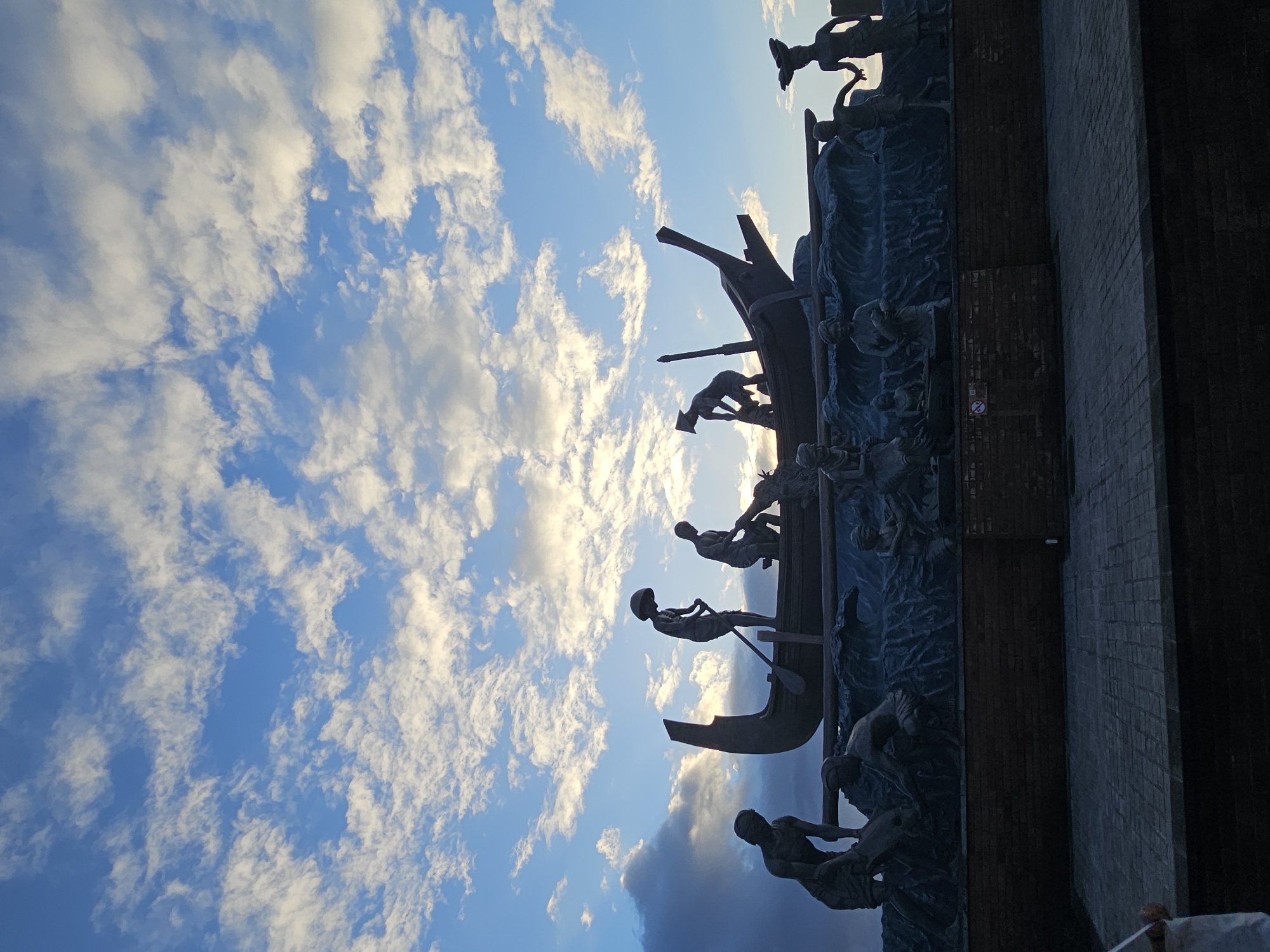 This image captures the "Bajra Sandhi Monument" in Denpasar, Bali, Indonesia. The monument is a large, silhouetted sculpture depicting figures in a boat amidst waves, with a backdrop of a dramatic sky filled with white clouds against a blue expanse. The sculpture represents the struggle and daily life of Balinese people, particularly fishermen. Several figures are visible in and around the boat, some rowing, others seemingly engaged in work. The monument is set on a raised brick platform, and in the foreground, a paved walkway can be seen. The lighting suggests it is either early morning or late afternoon, with the sun casting a warm glow on the clouds. The overall impression is one of strength and resilience conveyed through the artistic representation of human endeavor against the elements.