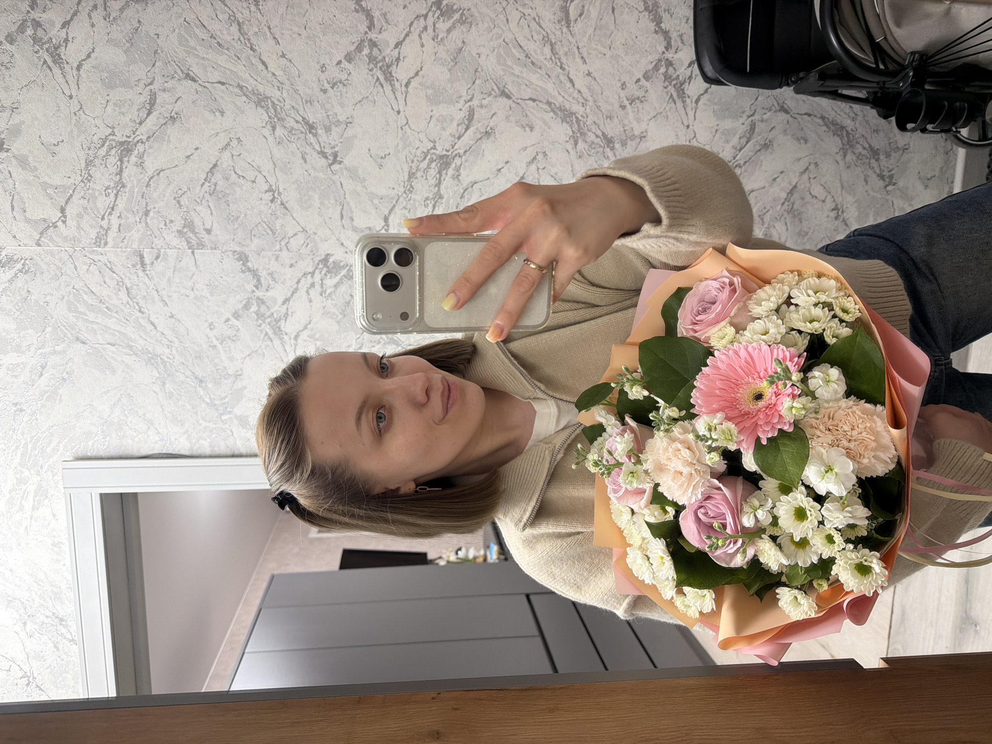 A woman is captured in a mirror selfie, holding a vibrant bouquet of flowers. She appears to be in her 20s or 30s, with light brown hair, blue eyes, and a neutral expression. She is wearing a light beige or cream-colored zip-up cardigan over a white top, paired with blue jeans. Her nails are painted light yellow, and she wears a simple gold-colored ring on her right ring finger.
She holds a silver-colored smartphone, likely an iPhone model with three camera lenses, encased in a clear, slightly sparkly protective case. The phone is used to take the selfie, with her arm extended.
The bouquet she holds is a mix of pink, white, and green flowers, wrapped in light peach and pink paper. Visible flowers include a prominent pink gerbera daisy, delicate light pink roses, clusters of small white chrysanthemums, and possibly light pink carnations, accompanied by various green leaves.
The background shows a wall covered in light gray and white marble-patterned wallpaper. Reflections in the mirror reveal further details of the indoor setting. In the upper right, a dark object resembling part of a stroller or a wheeled chair is visible. In the lower left, a light-colored doorway and a modern dark gray cabinet or counter surface are reflected. A wooden surface, possibly a mirror frame or furniture, frames the bottom edge of the image. The scene suggests a personal indoor space, likely a home or apartment in Reshetnikovo, Russia, during daytime. There is no visible text in the image.