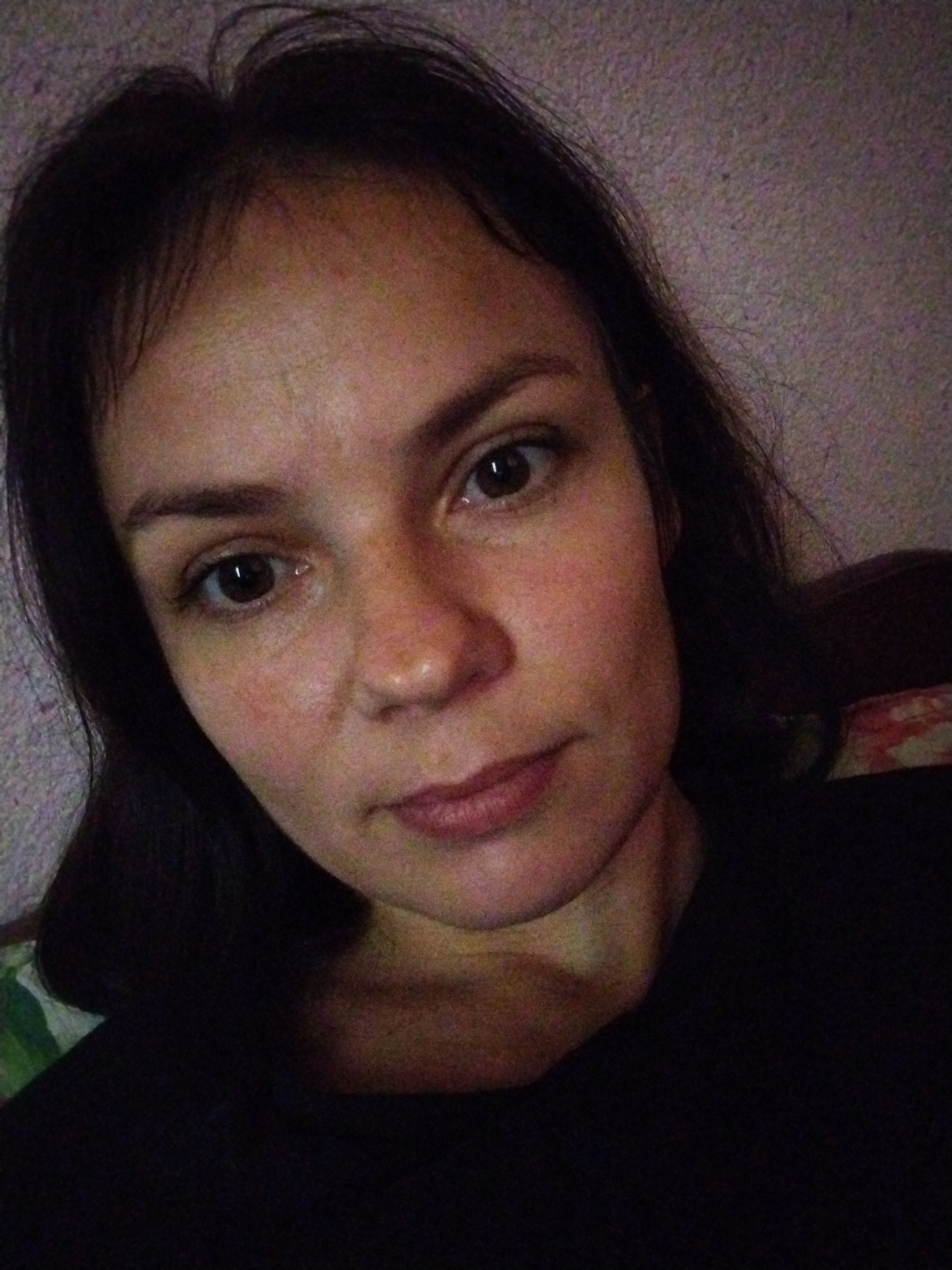 A close-up, front-facing selfie captures a woman with dark hair and fair skin. Her head is tilted slightly to her right. Her eyes are open and looking towards the camera. The background is a textured, light-colored wall. The lighting is dim and appears to be from an artificial source. The woman is wearing a dark-colored top. No specific location in Molodiia, Ukraine can be determined from the image.