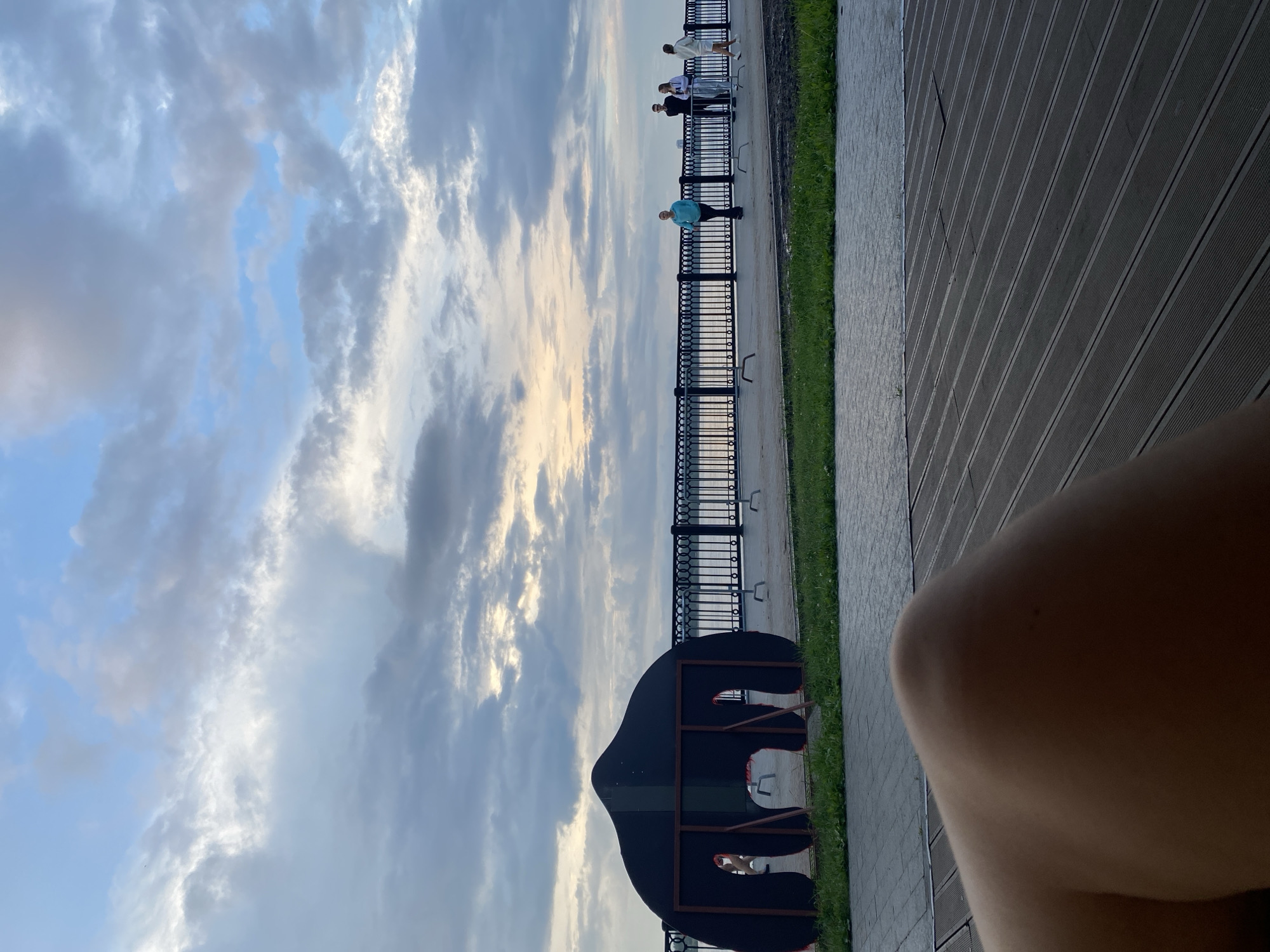 The image depicts a waterfront promenade with a cloudy sky overhead. In the foreground, a person's leg and knee are visible, resting on a wooden deck. To the left, a large, dark, abstract sculpture resembling an elephant with openings is present, situated on grass. Beyond the sculpture, a metal railing lines the promenade. Several individuals are seen walking and standing along the promenade, facing the water. The water appears calm, and the sky is a mix of blue and grey clouds, with hints of sunlight breaking through. The location is the Embankment in the city of Perm, Russia, likely near the Kama River.