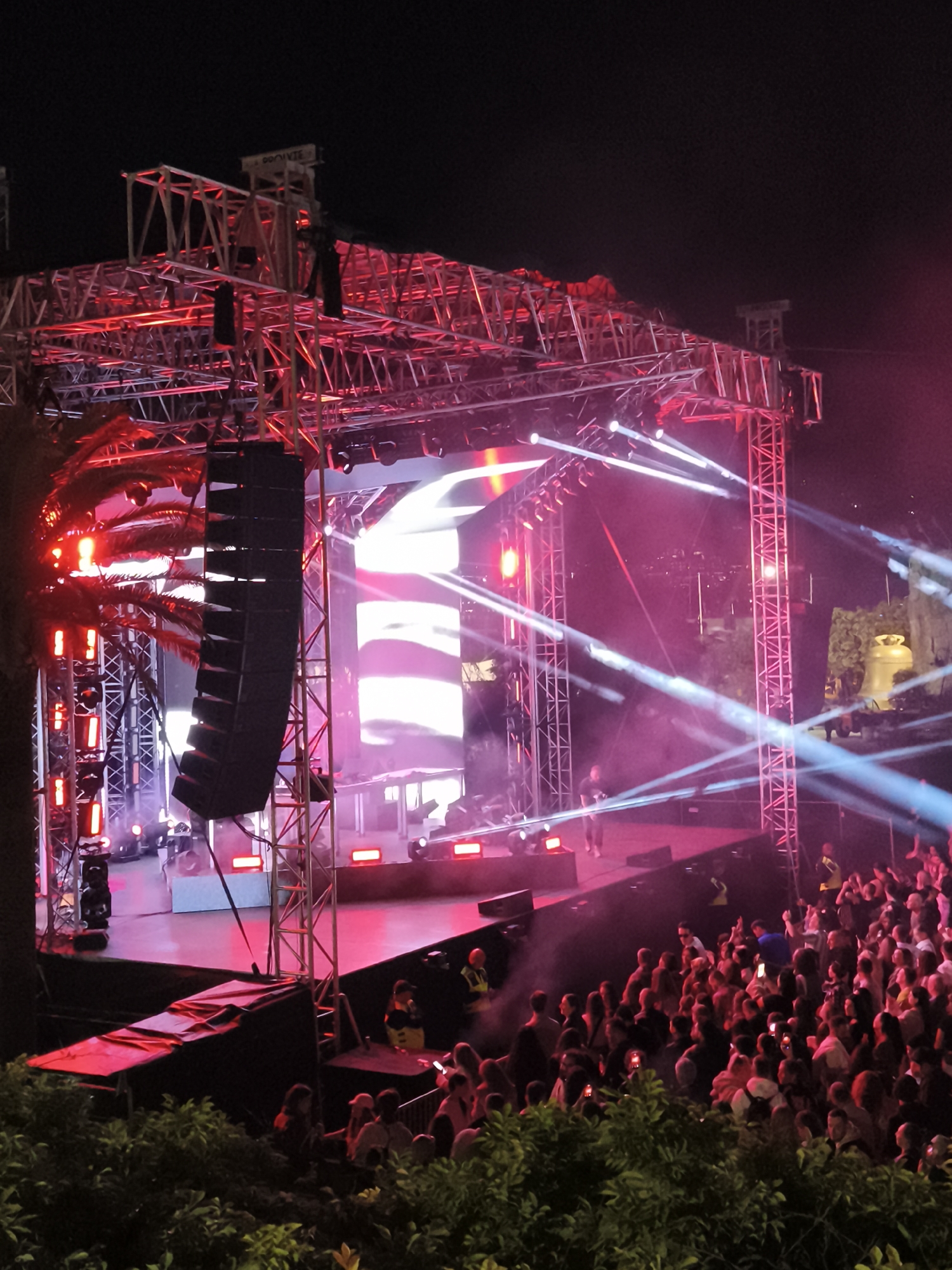 A nighttime concert scene is captured from a low angle, seemingly from behind shrubbery or low bushes. The stage is a complex structure of metal scaffolding, illuminated with red and white lights. A large vertical speaker array is visible on the left side of the stage. A performer, partially obscured by light and distance, stands on the stage, lit by a variety of stage lights. Beams of white light cut through the air. A crowd of people faces the stage, many holding up phones to record or photograph the event. The lighting is primarily red and white. The location is recognizable as near the ancient bell tower and the Citadel in Budva, Montenegro, judging by the structures visible amidst the lights.