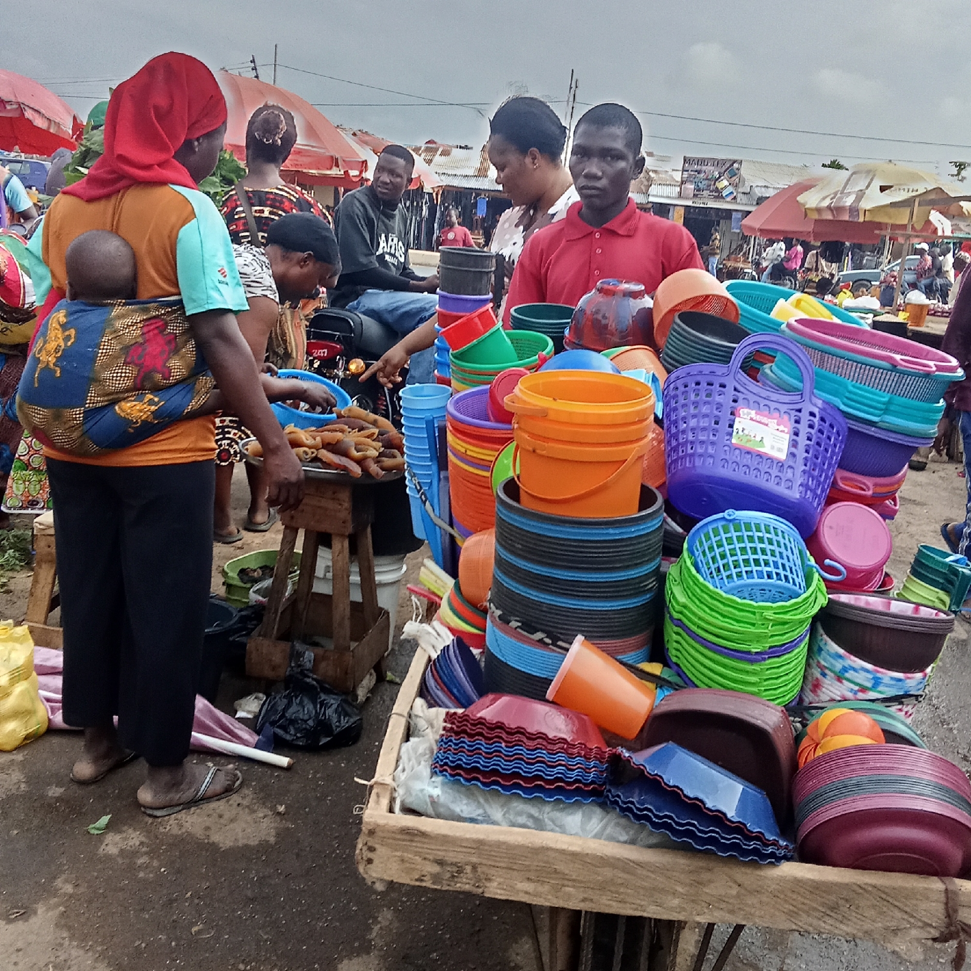 The image depicts a market scene in Akwanga, Nigeria. A vendor wearing a red shirt stands behind a wooden cart heavily loaded with stacks of colorful plastic buckets, baskets, and bowls. To the left, a woman wearing a red head covering, an orange top, and carrying a baby wrapped on her back is looking at food items on a small table. Another woman is also looking at the items. A man sits on a motorcycle nearby. The market has vendors underneath umbrellas and cloudy skies overhead.