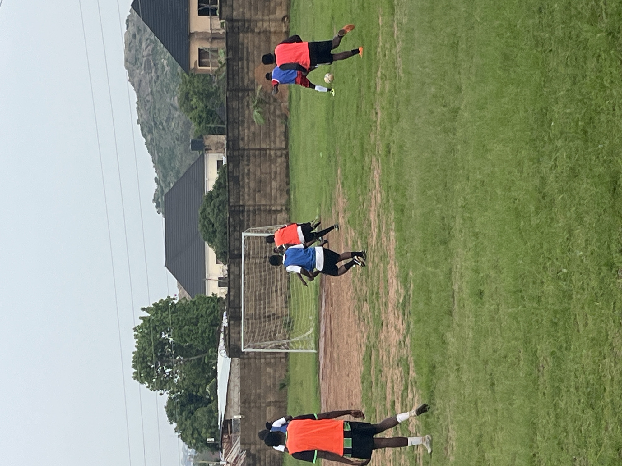 The image depicts an outdoor soccer training session on a grassy field in Jos, Nigeria. Several individuals, primarily young men, are engaged in the activity. They are dressed in athletic wear, including brightly colored jerseys and shorts. Two individuals in the foreground are wearing orange and black, while others are in blue and red. One person is actively kicking a soccer ball, suggesting a practice drill or game is in progress. In the background, a soccer goal is visible, further confirming the context.
The setting includes elements of a semi-urban environment. Buildings, some with unfinished construction, and a brick wall are visible behind the field. A prominent rocky hill formation and lush green trees are also present, indicating the natural landscape surrounding the training area. The sky appears overcast, suggesting it might be a cloudy day, possibly in the morning or late afternoon given the soft lighting. There is no visible text in the image. The overall atmosphere appears to be one of focused athletic practice.