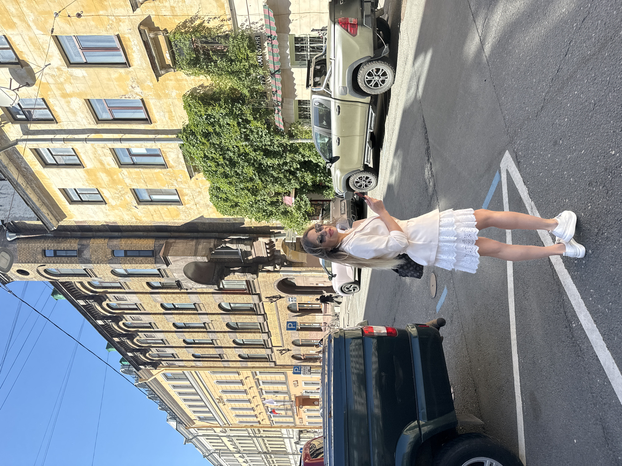 A woman stands on a street in Saint Petersburg, Russia, holding a mobile phone and looking at the camera. She is wearing a white dress with lace trim and white sneakers. The street is paved with asphalt and marked with white lines. To her left, a dark green van is parked. Behind the van, a beige pickup truck is visible. Further back, an old, yellow brick building with many windows and ornate architectural details lines the street. Green foliage covers part of the building's facade. The scene suggests a sunny day, likely daytime, given the bright sunlight casting shadows. The woman's expression is neutral, and the overall atmosphere appears casual and urban. No visible text is legible in the image.